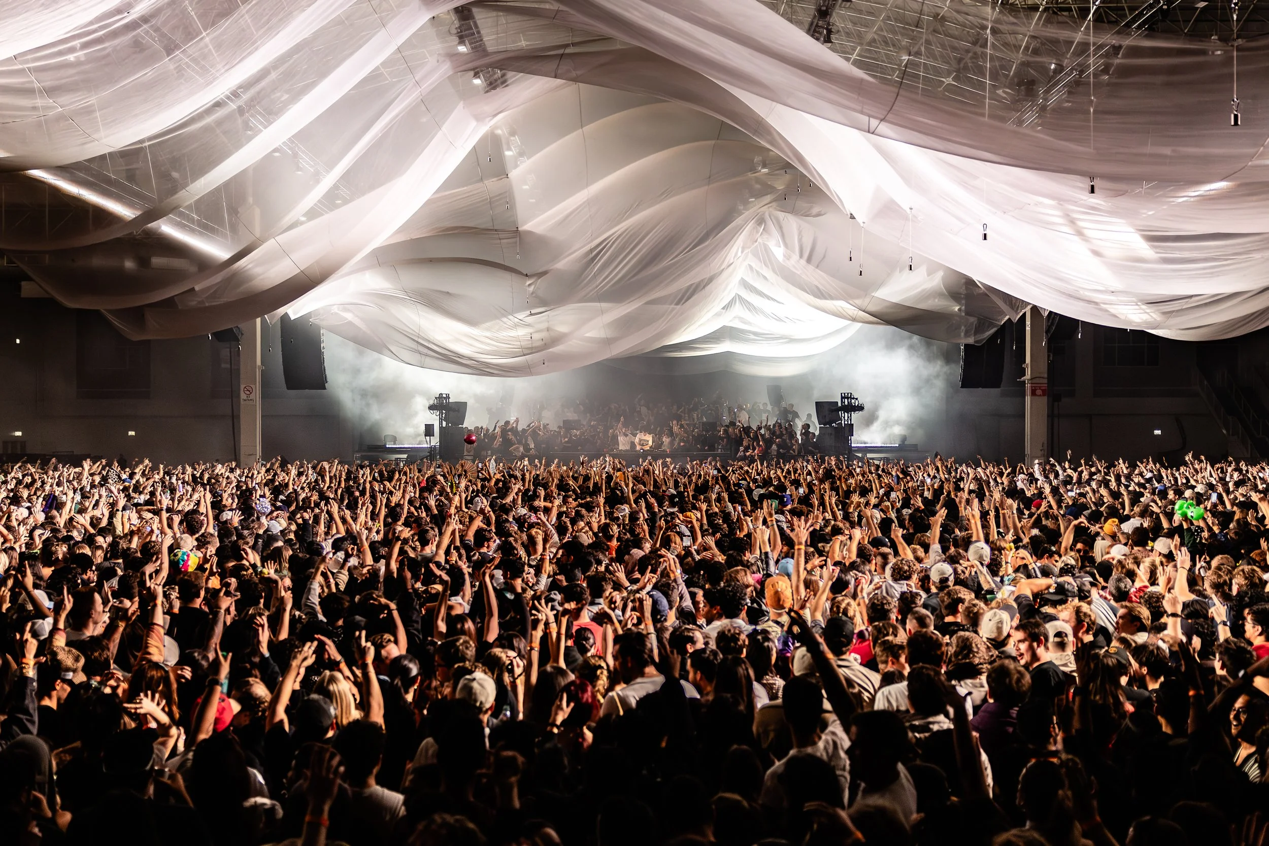 Large indoor concert venue filled with a crowd of people raising their hands, facing a stage with performers and sound equipment, under draped white fabric ceiling.