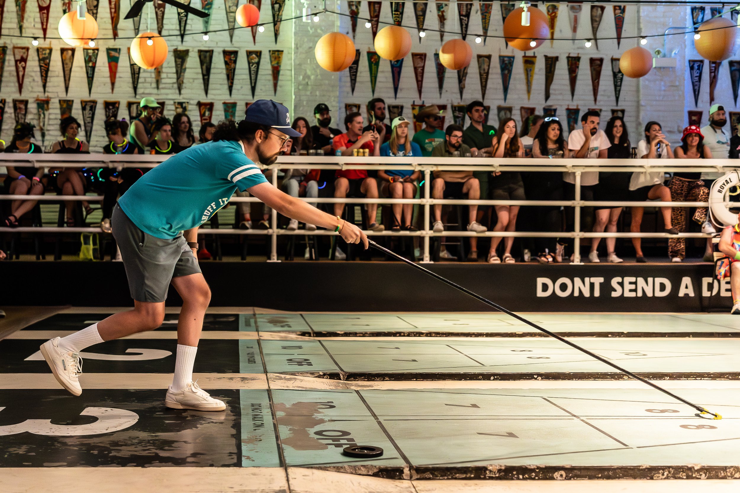A young man in casual sportswear is playing shuffleboard on an indoor court, with a large audience seated behind him and decorative hanging lanterns and banners overhead.