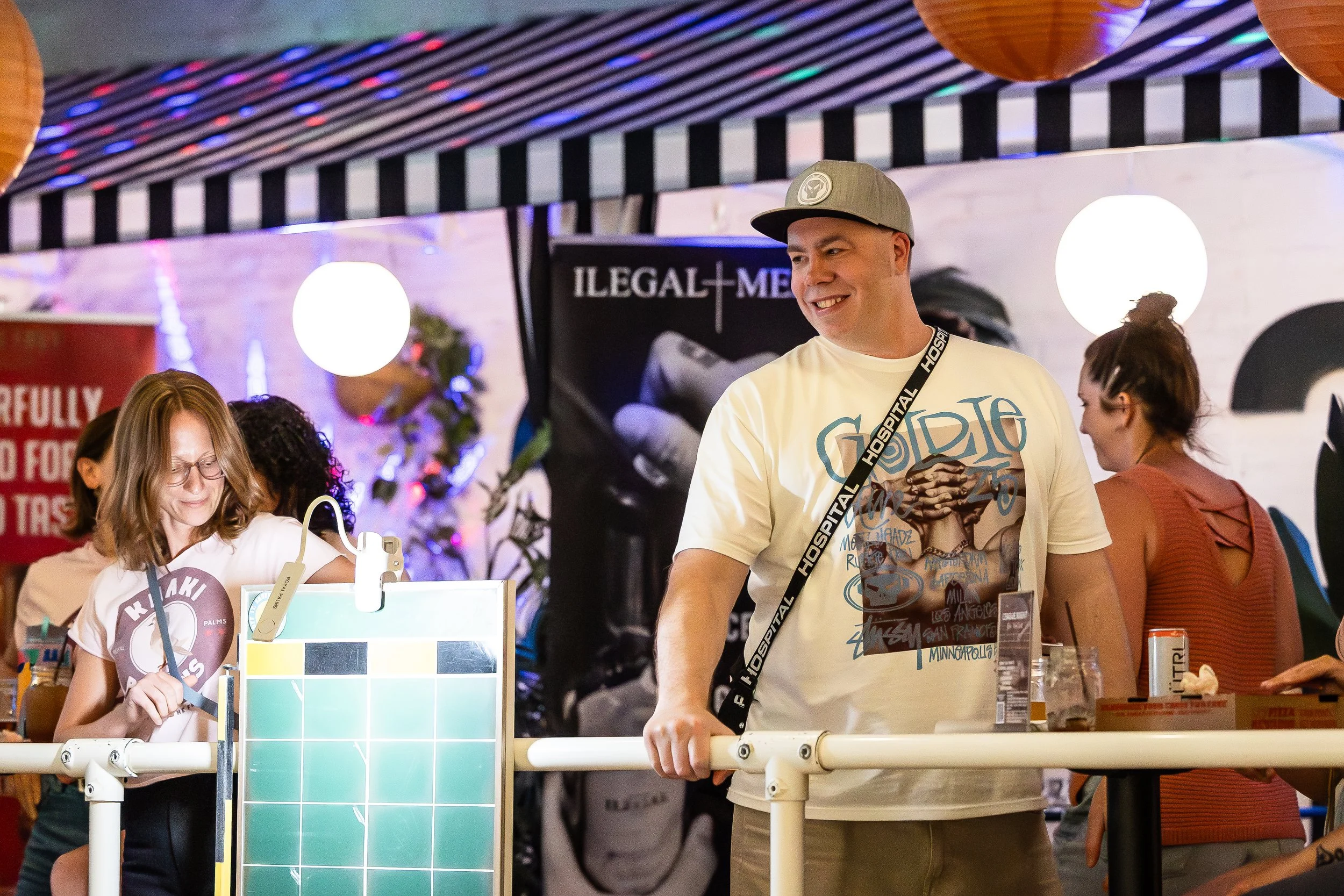 People socializing at an indoor event, with a woman wearing glasses and a graphic t-shirt holding a pen, and a man in a gray cap and a graphic t-shirt smiling and standing near a railing.