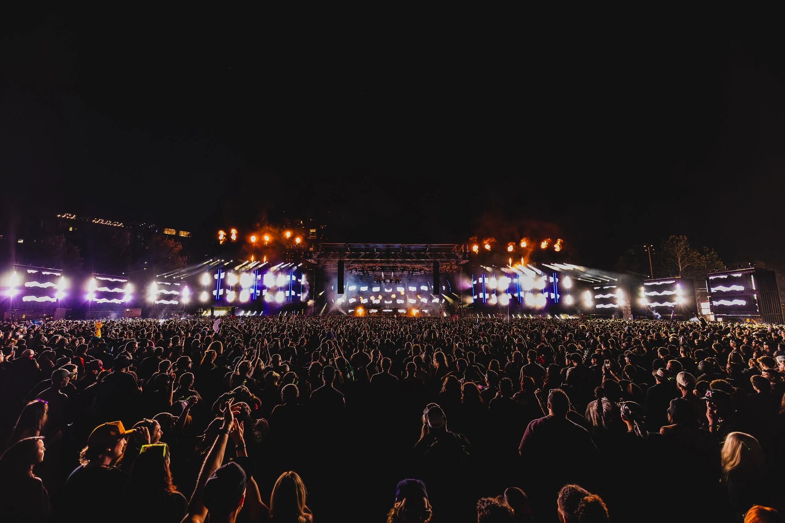 Nighttime outdoor concert with a large crowd facing a brightly lit stage with smoke and pyrotechnics.