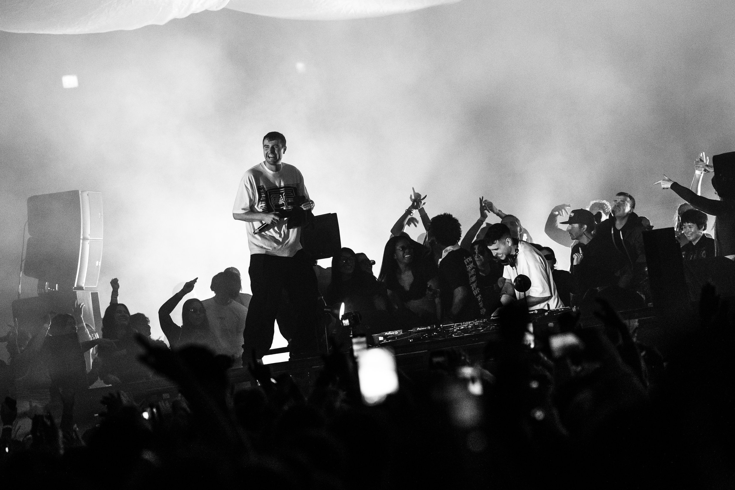 Crowd of people dancing and enjoying music at a concert or event, with Fred Again standing on stage in front of speakers, in black and white.