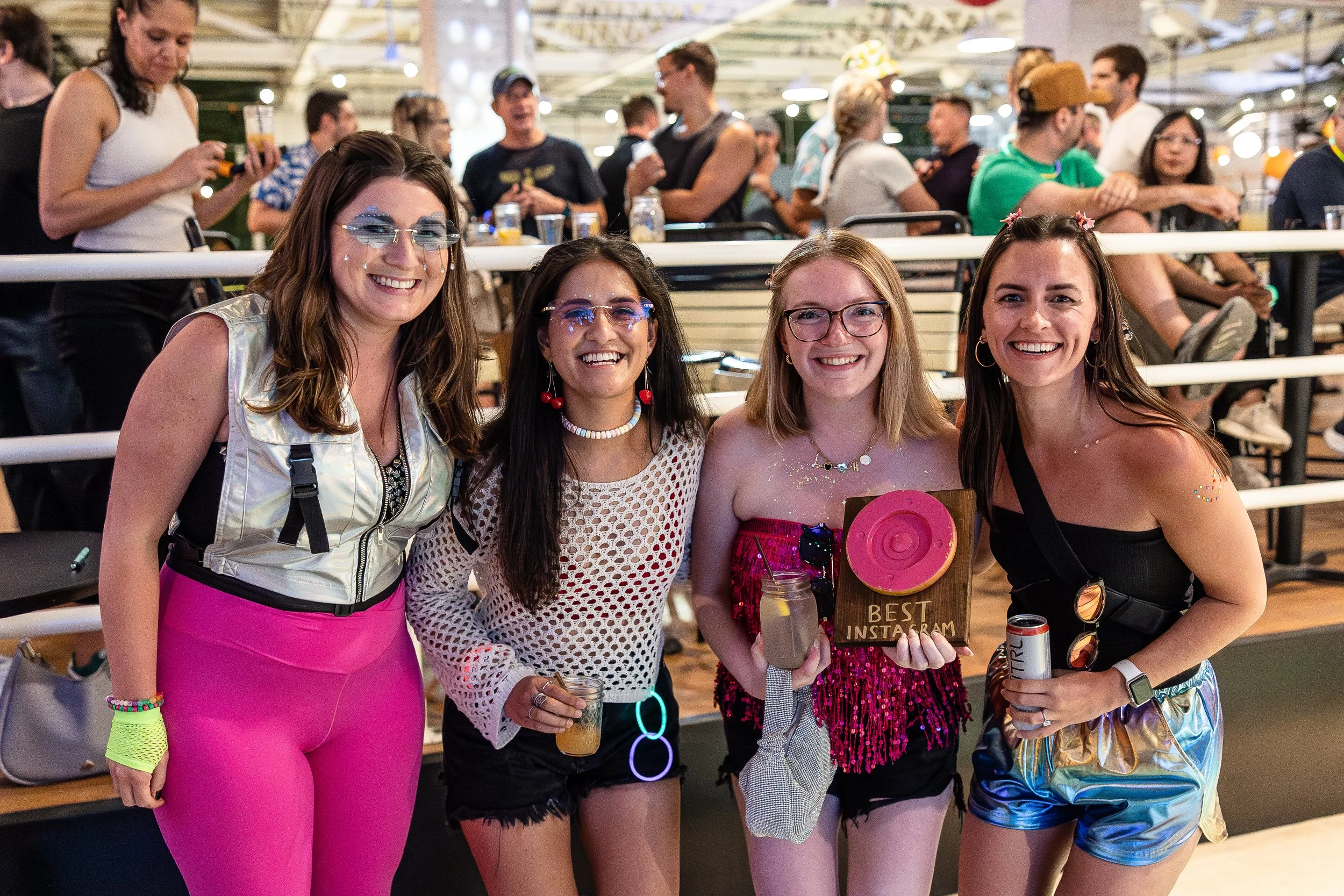 Four women celebrating at a party, holding drinks and a wooden plaque that reads 'Best Instagram,' with a crowd of people in the background.