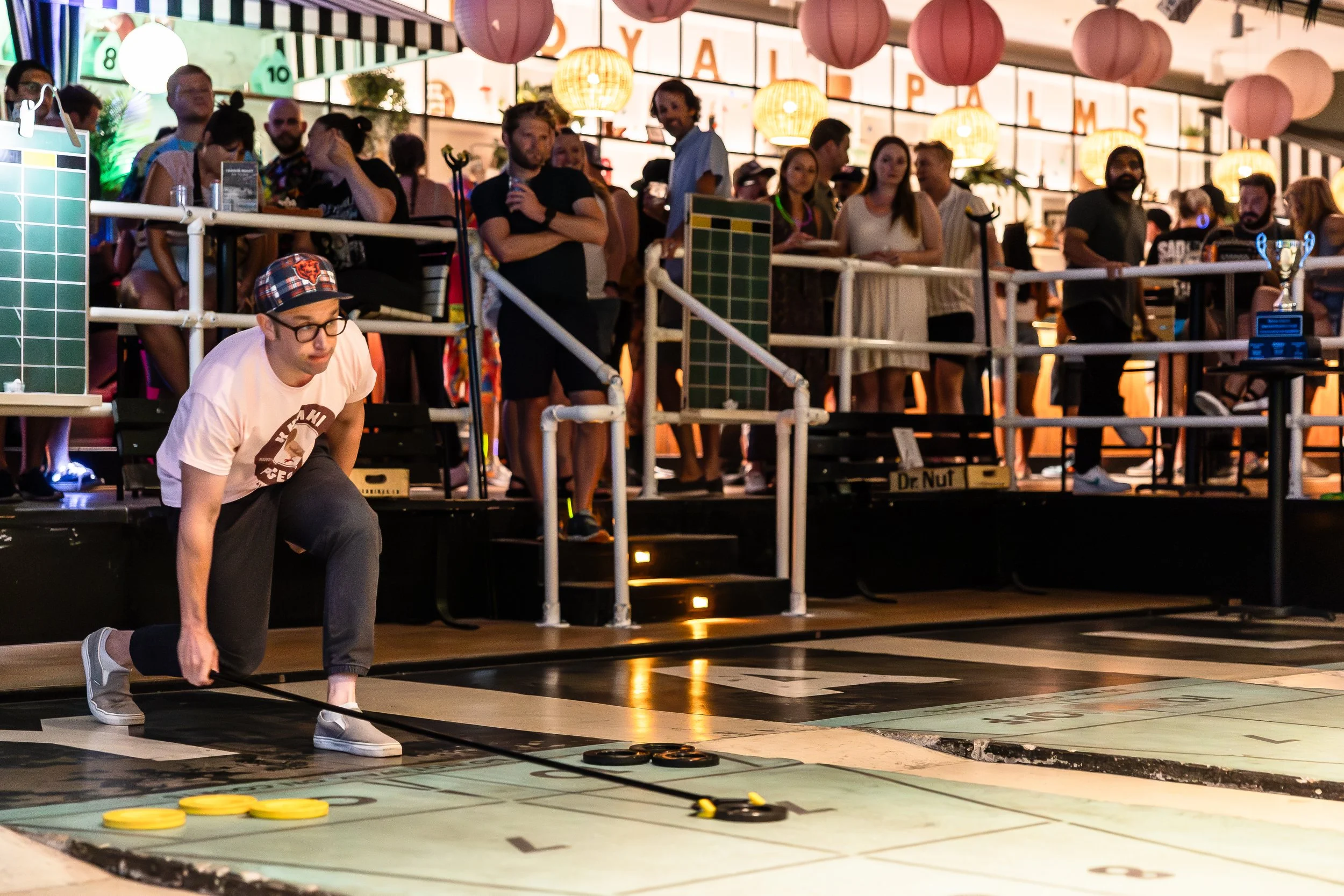 A man in a white t-shirt, glasses, and a plaid cap bends down to pick up disks on a shuffleboard court, with a crowd of spectators watching from behind a railing in a lively indoor setting decorated with pink lanterns and illuminated signs.