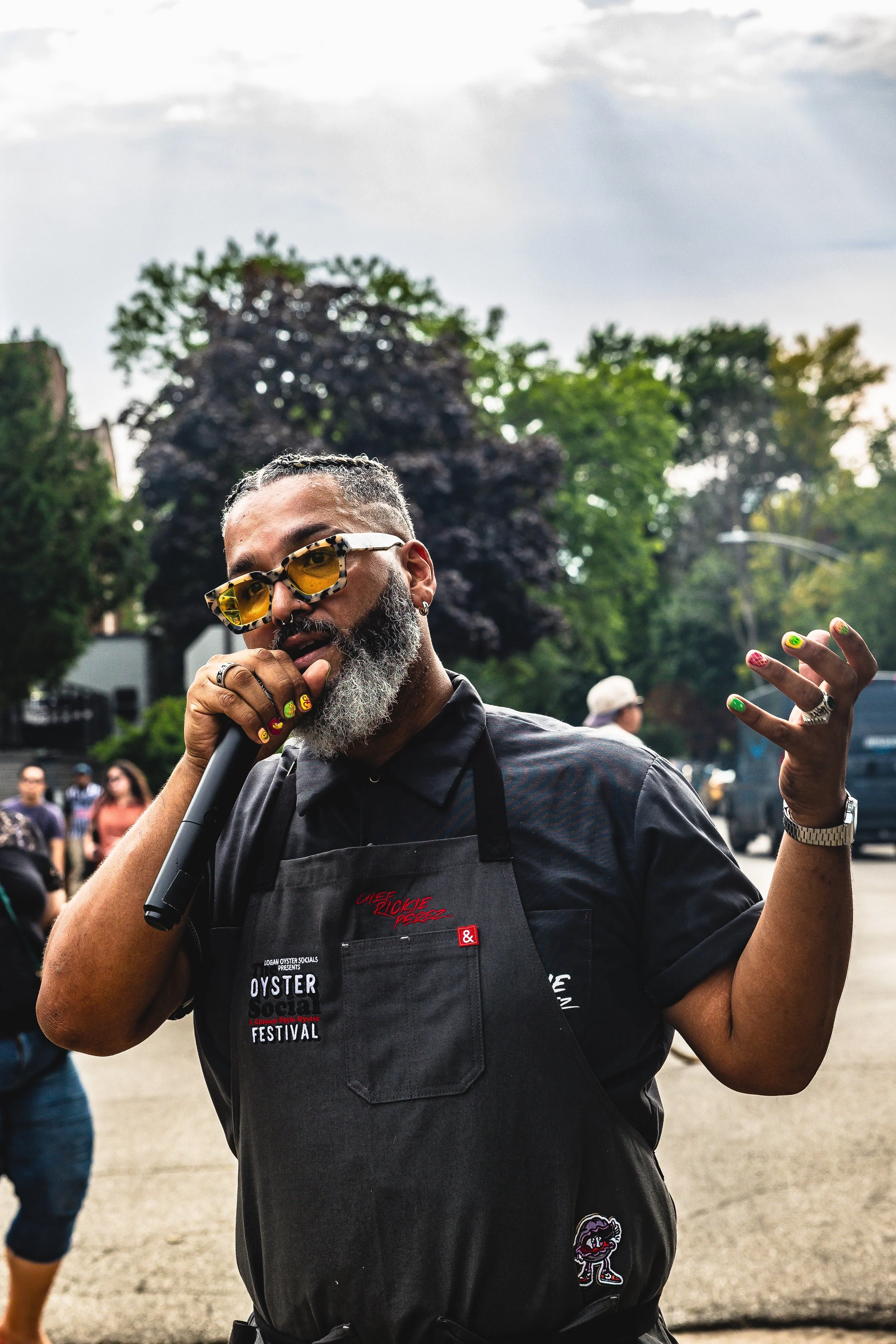Man with gray beard wearing sunglasses, black shirt, and apron at an outdoor event, holding a microphone, with trees and people in the background.