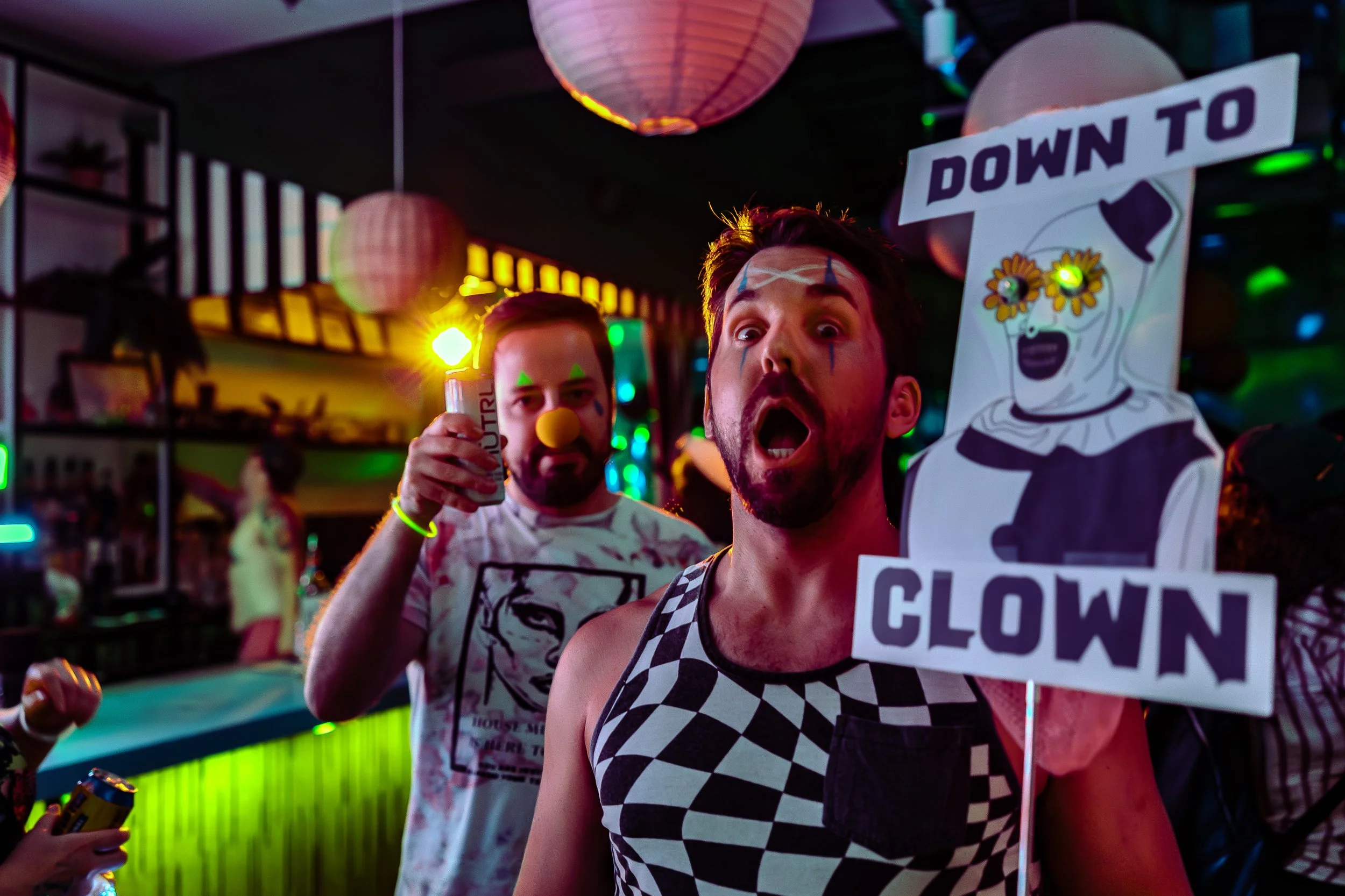 Two men dressed in clown costumes at a lively party, one holding a sign that says 'Down to Clown' and wearing a checkered tank top, with colorful lights and lanterns in the background.