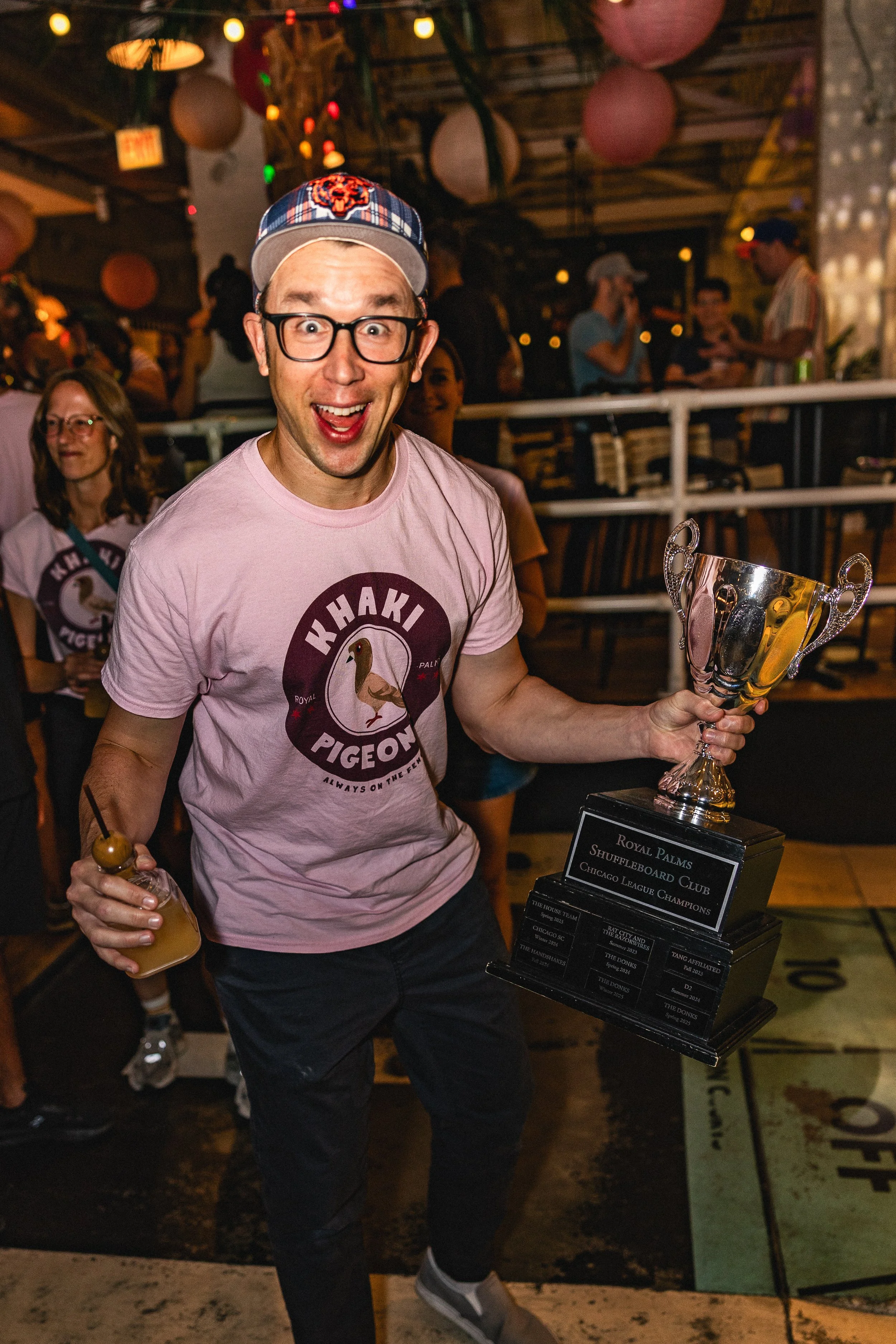 A man wearing glasses and a Chicago Bears hat holding a trophy in one hand and a drink in the other at a celebration event. There are people in the background enjoying the party.