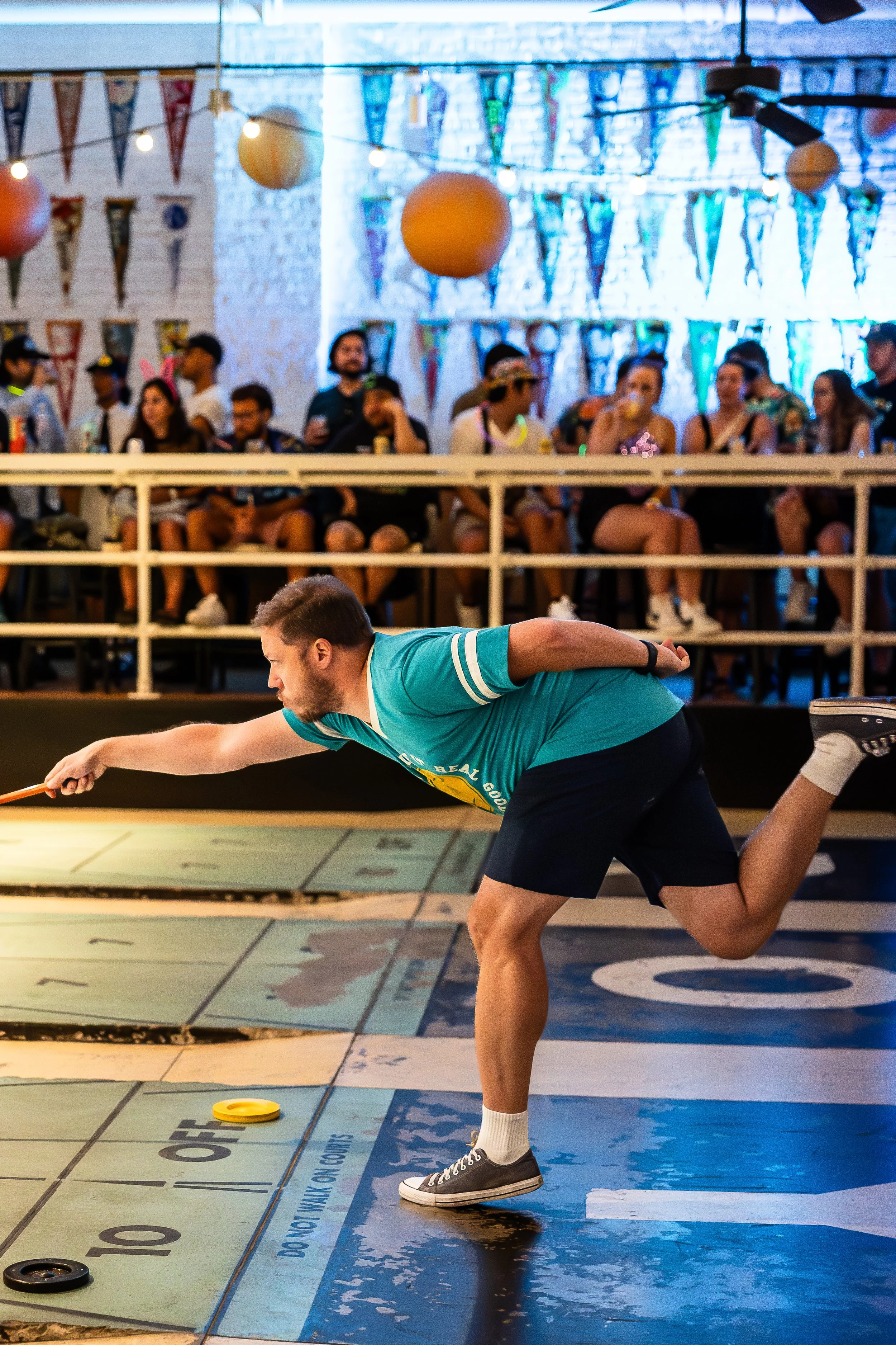A man playing shuffleboard indoors, reaching to slide a disc on the court, with an audience watching from behind a barrier.