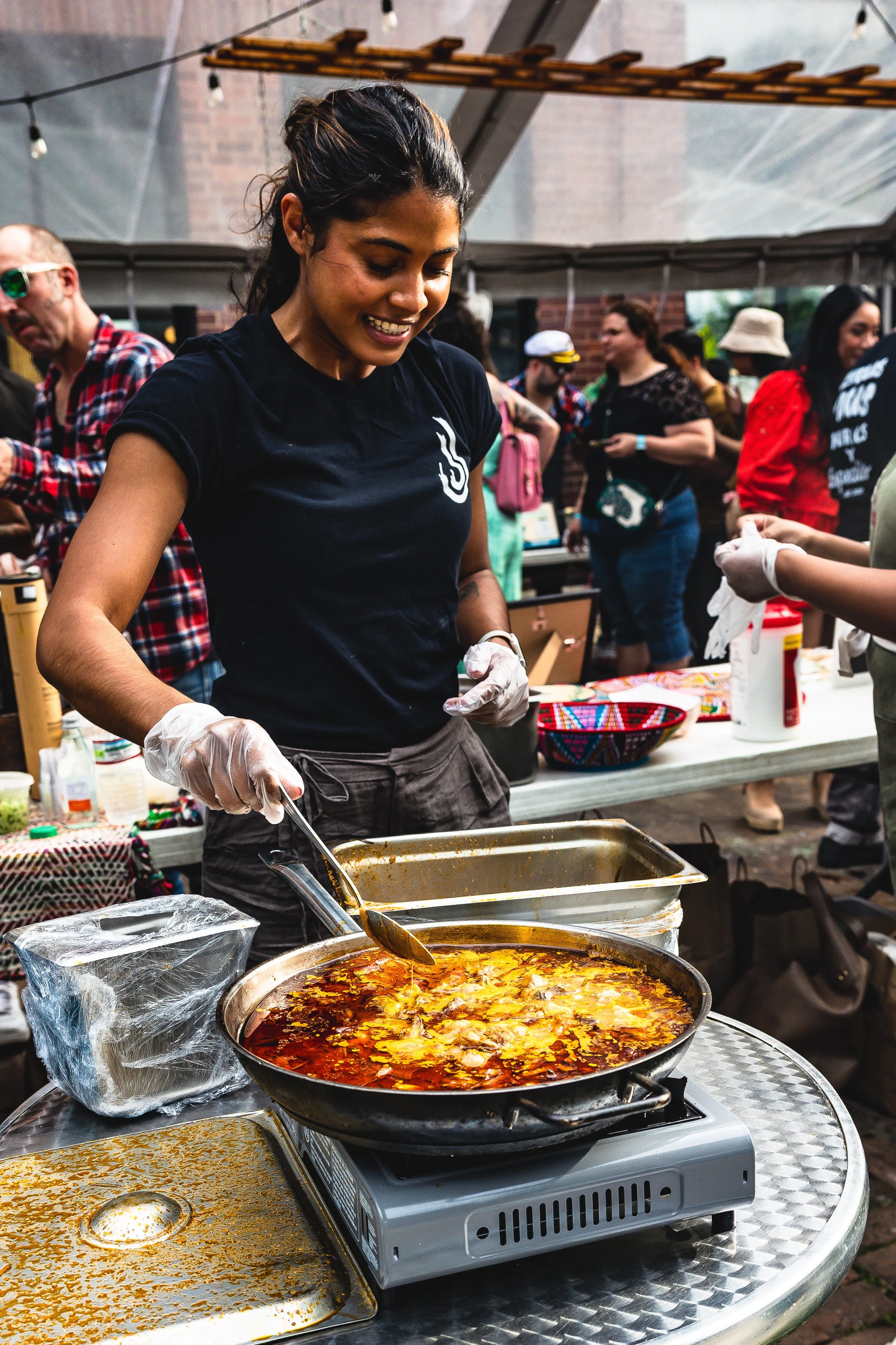 A woman cooking food at an outdoor food stall during a busy event or festival. She is wearing a black T-shirt, gloves, and is stirring a large pan of simmering stew or soup with a ladle. Several people are visible in the background under a canopy, so