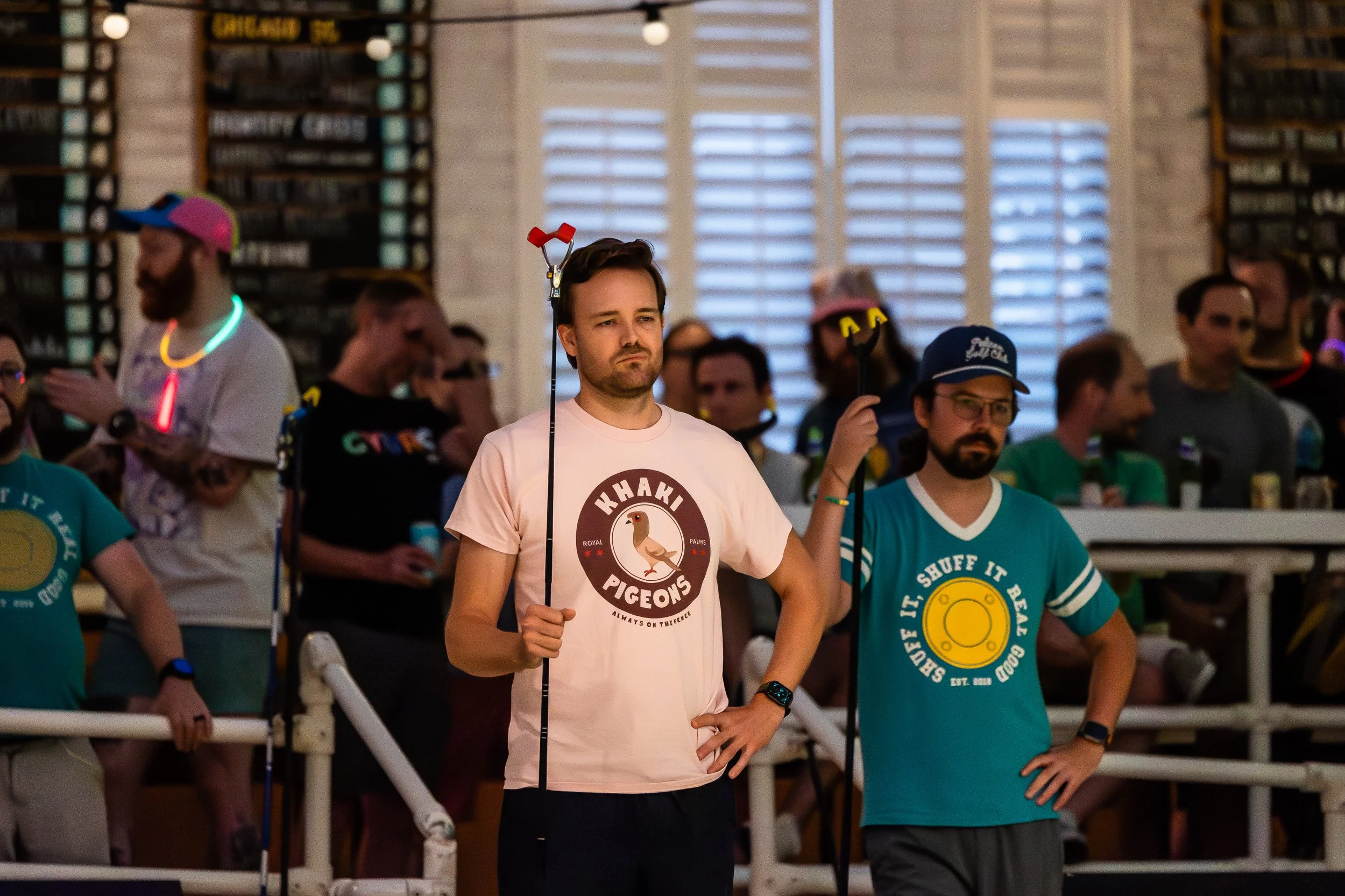 Group of people at a lively indoor event, some wearing glow-in-the-dark accessories and holographic wear, with two men in the foreground holding hockey sticks. One wears a pink shirt with a pigeon graphic and the other a teal shirt with a yellow disc