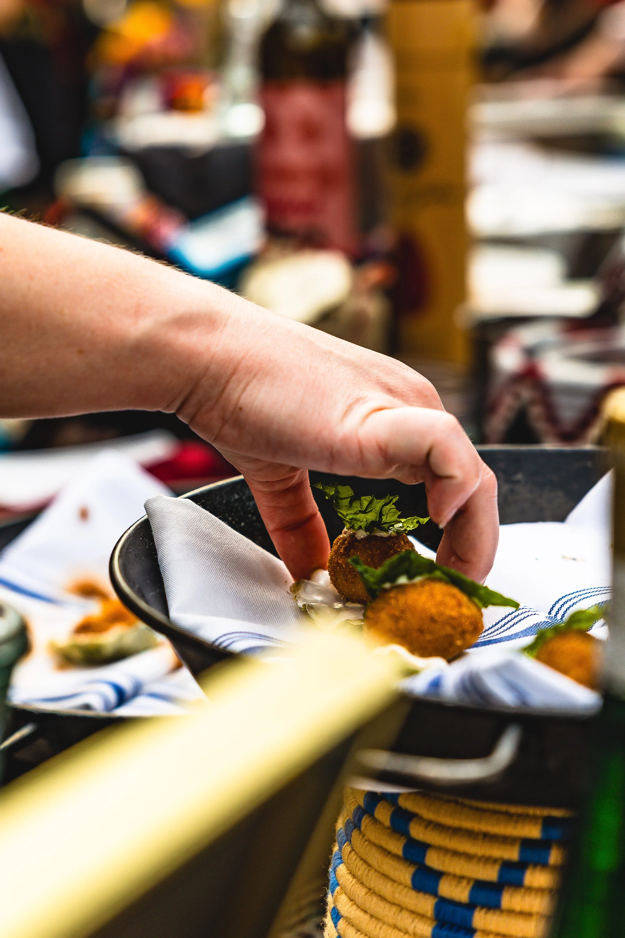 A hand reaching to pick up fried appetizers garnished with leafy greens on a black plate lined with white and blue cloth at a food market or restaurant.