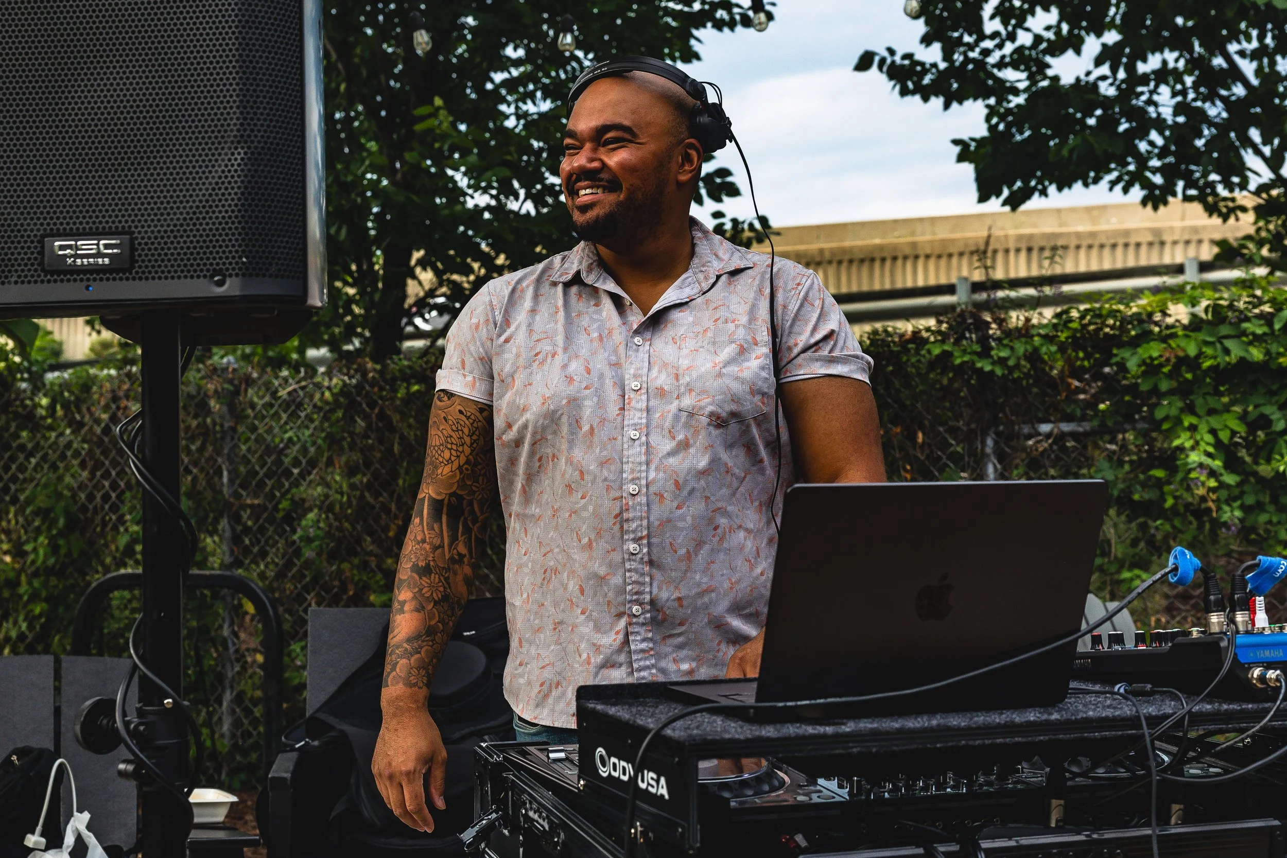 A man with a tattooed arm wearing a short-sleeved, patterned button-up shirt is DJing outdoors, smiling and wearing headphones, with a sound system and laptop in front of him, and trees and a fence in the background.