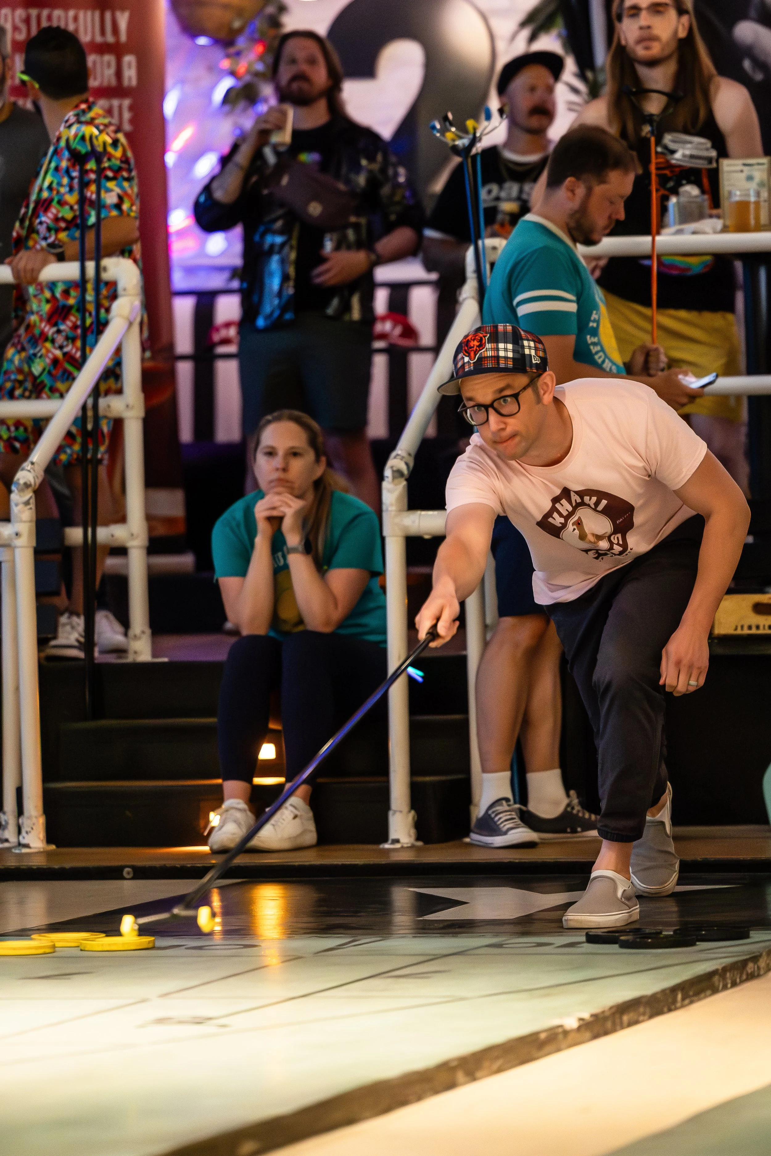 A group of people watching a man playing shuffleboard in an indoor arcade or entertainment center.