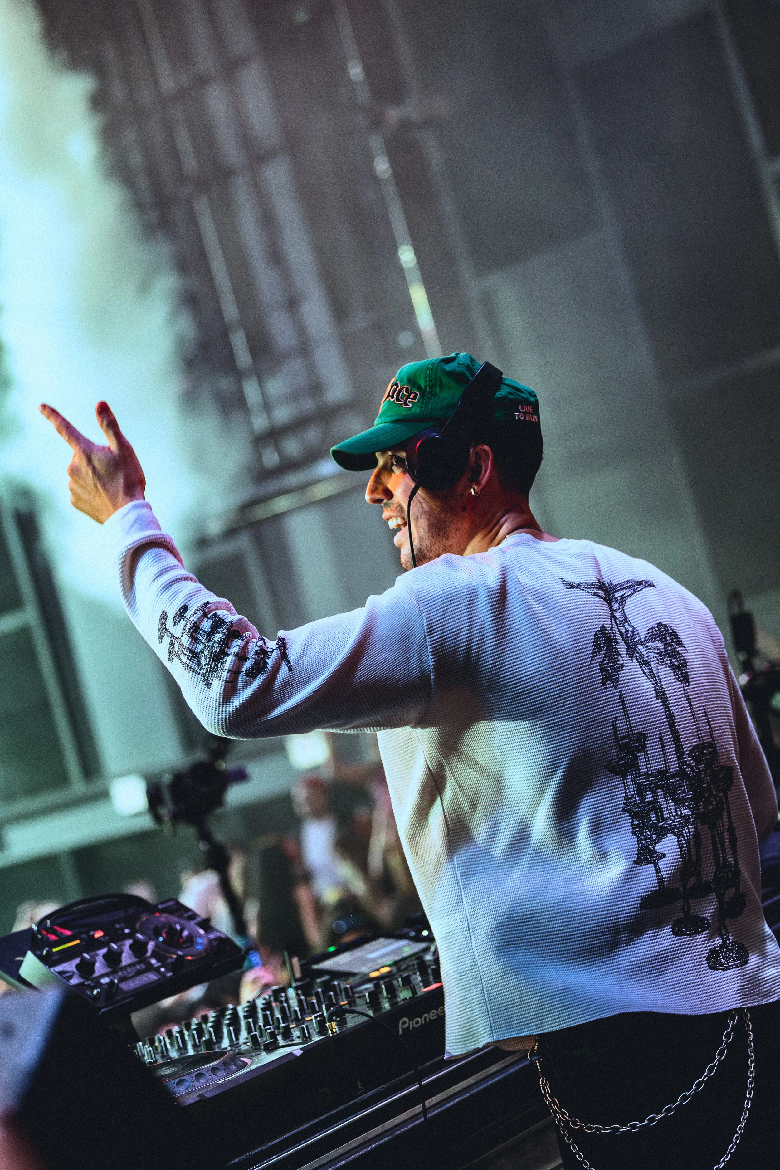 Cloonee wearing a green cap and white sweater with black palm tree sketches, smiling while operating DJ equipment at a club or music event.
