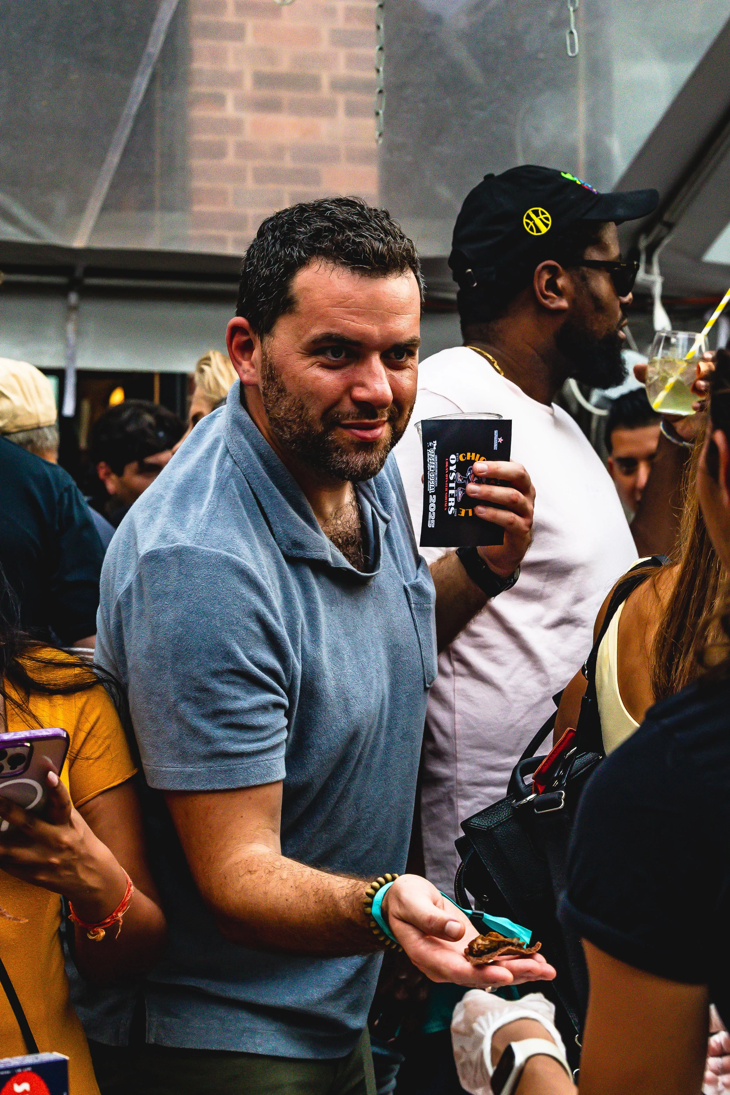 A man with a beard and short curly hair wearing a gray polo shirt, holding a cookie in one hand and a ticket or card in the other, stands in a crowded room at an event or party.