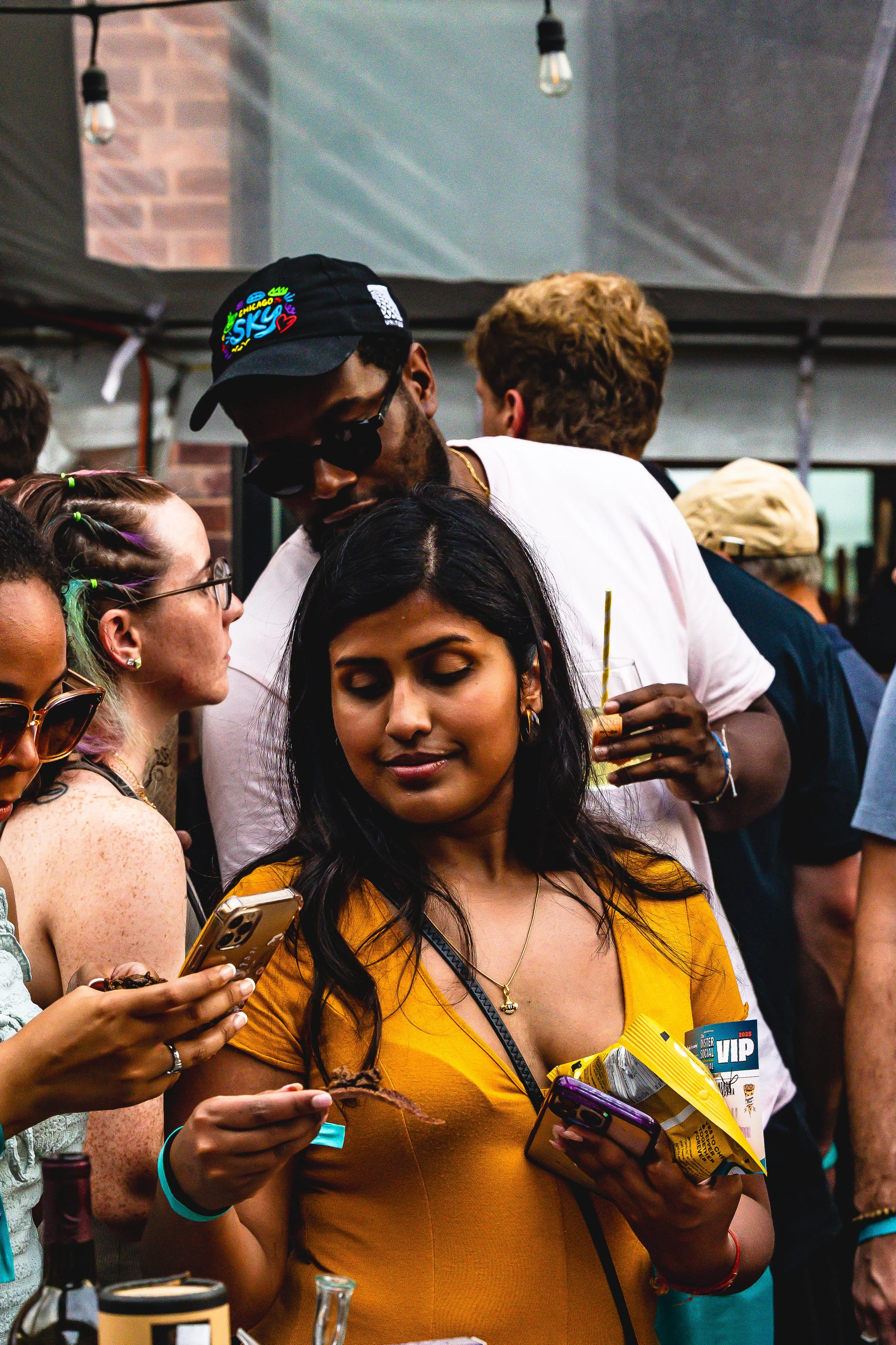 Group of diverse people socializing at an outdoor event, some are looking at their phones, with drinks in hand, under string lights on a covered patio.