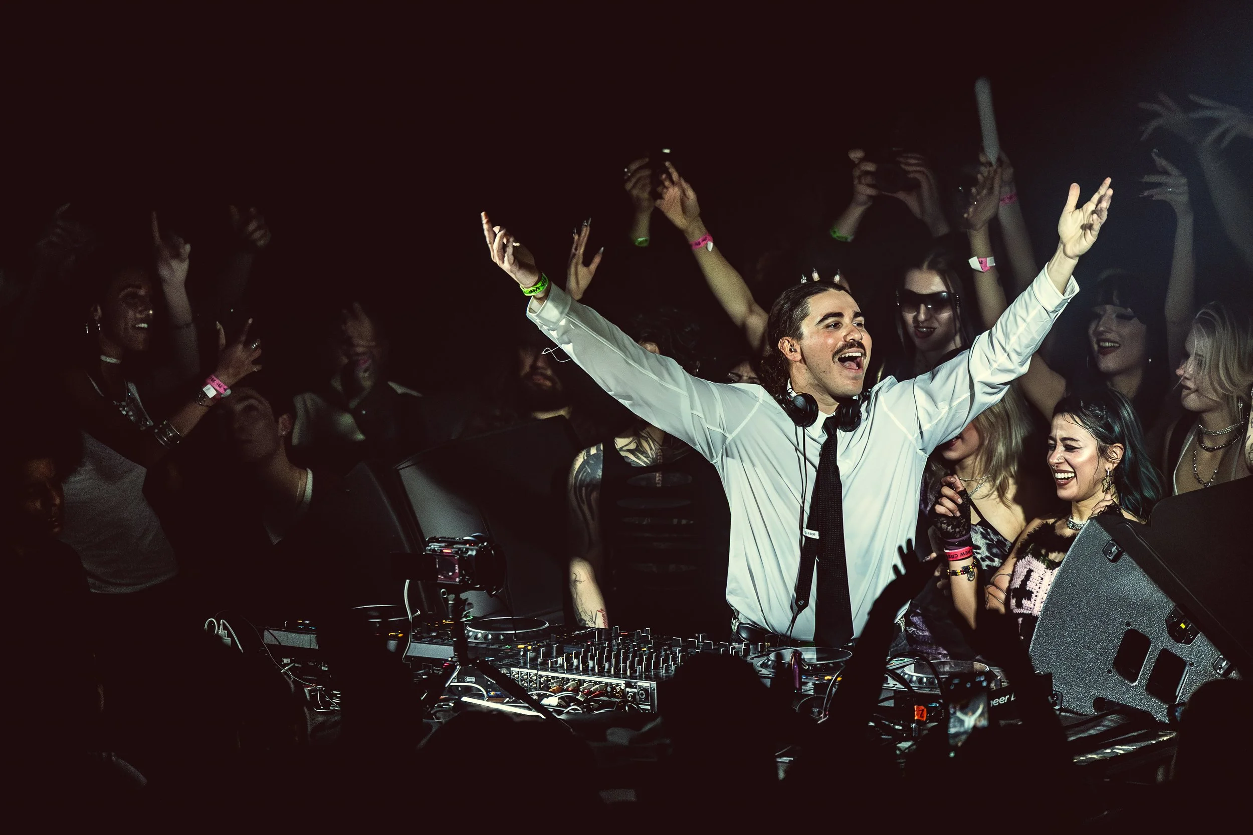 HHunter, a DJ with long hair and a mustache, wearing a white shirt and a black tie, is performing at a crowded nightclub. He is smiling with his arms raised, surrounded by cheering crowd members dancing and enjoying the music.