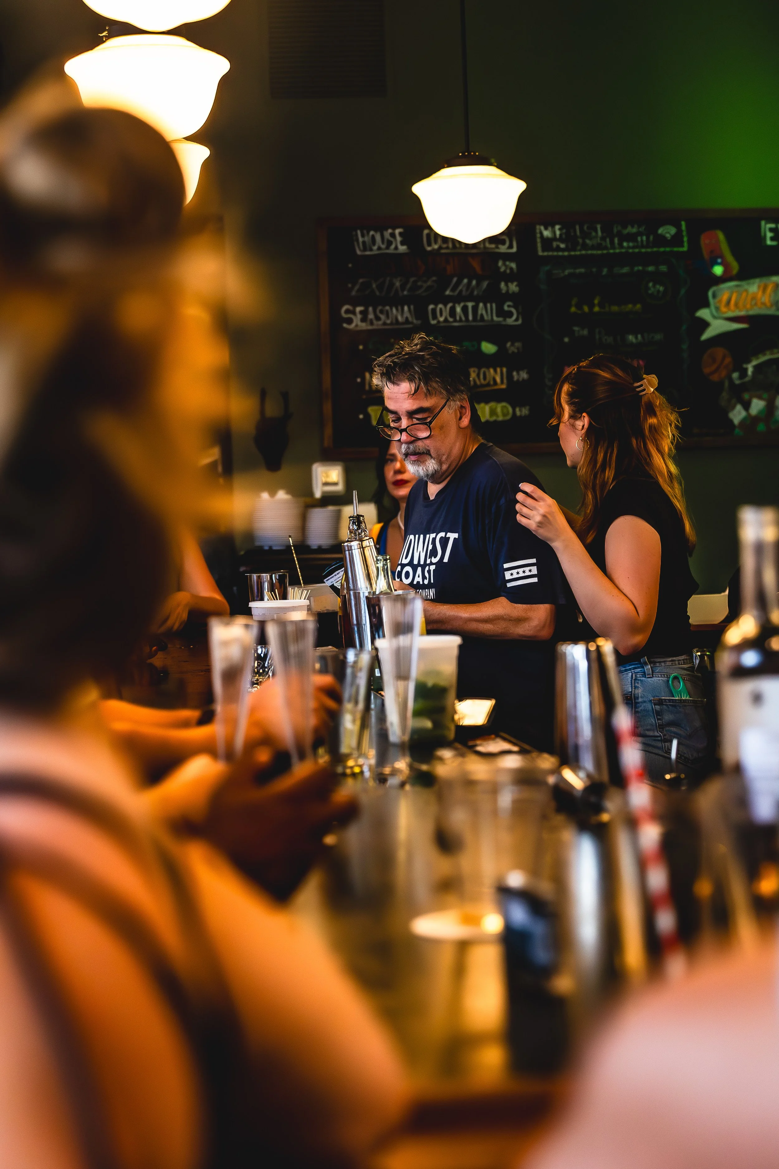 A bartender preparing drinks behind a bar with customers sitting in front, in a dimly lit tavern or restaurant with hanging lights and a chalkboard menu in the background.