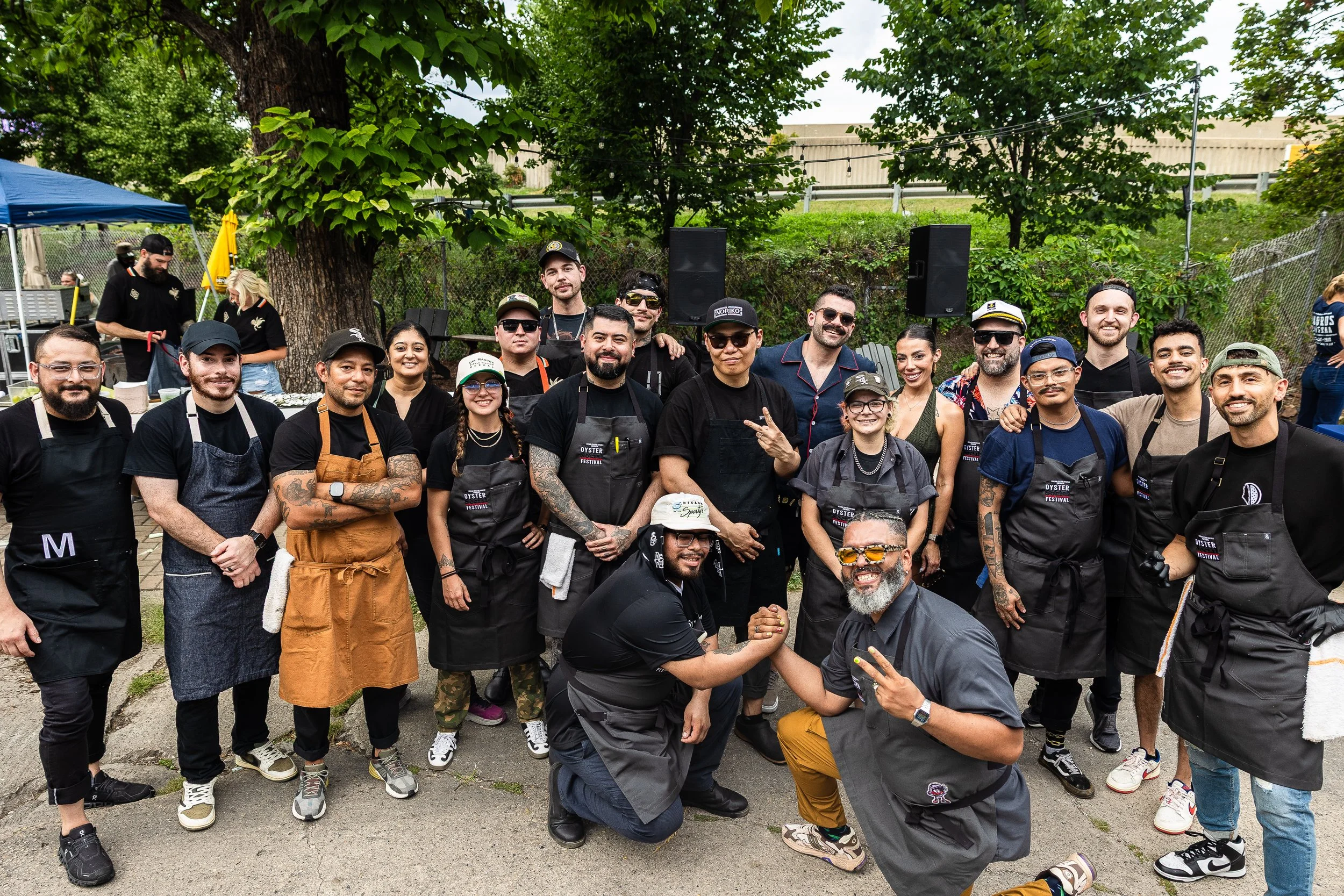 Group of people outdoors at a festival or gathering, some wearing aprons, standing in front of a tree and speakers, smiling and posing for the photo.