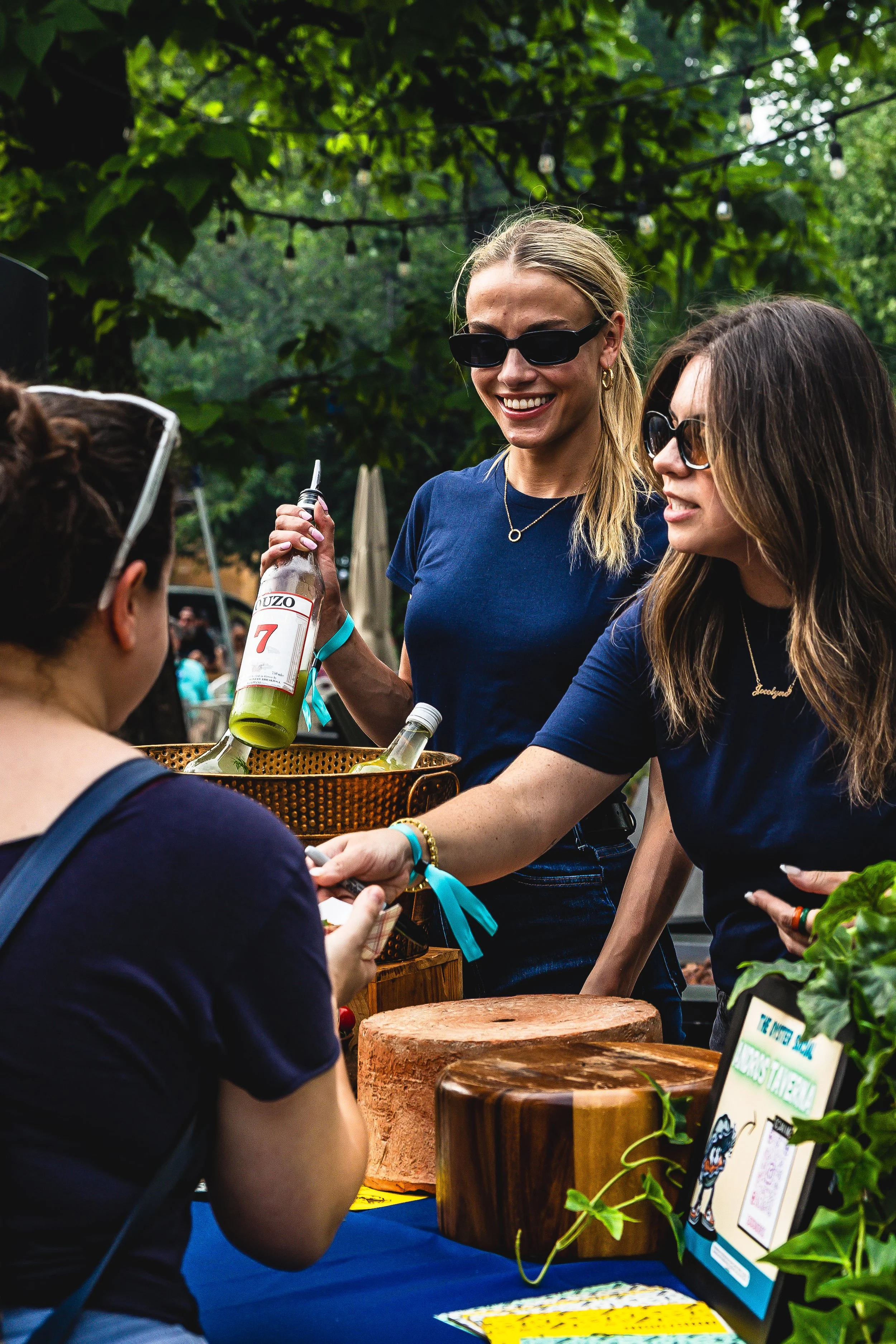 Two women serving drinks at an outdoor event or market, with a woman receiving a drink. All are wearing sunglasses and smiling, with trees and string lights overhead.