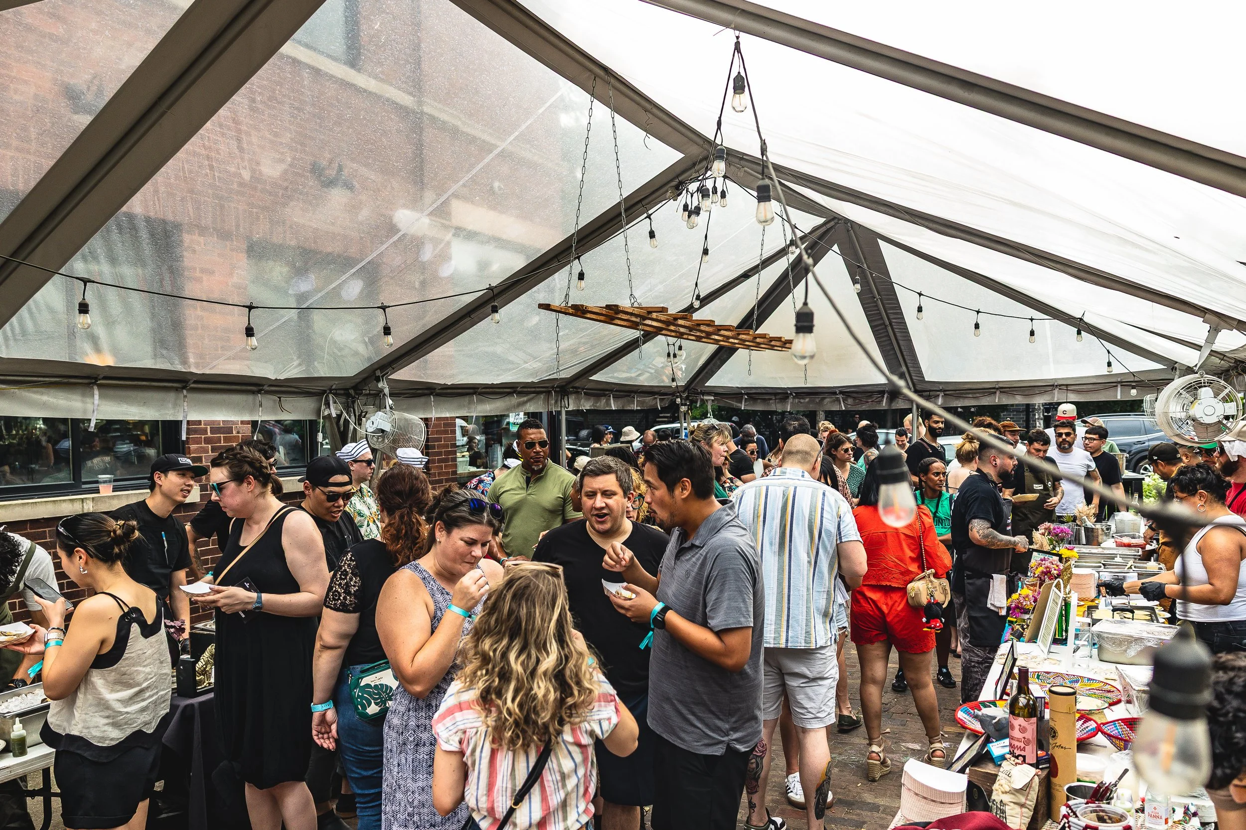 Crowd of people at an outdoor event under a large tent, socializing and browsing tables with items for sale or display.