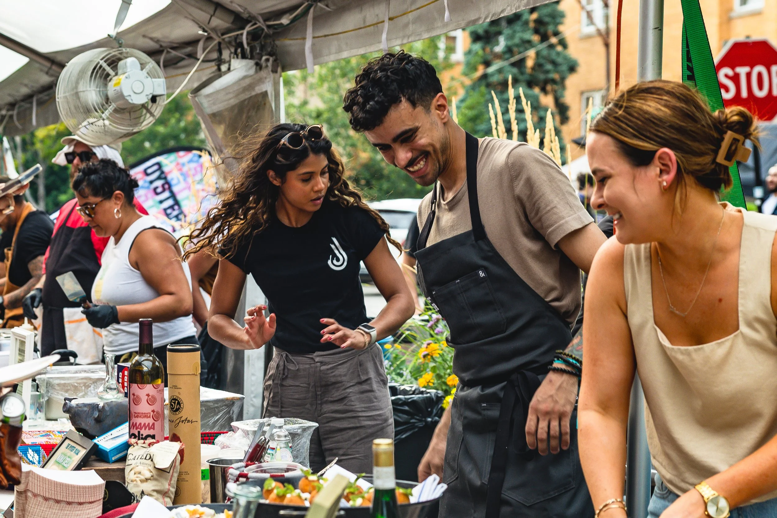 People smiling and interacting at an outdoor market stall with various food items and beverages, under a canopy with a fan.