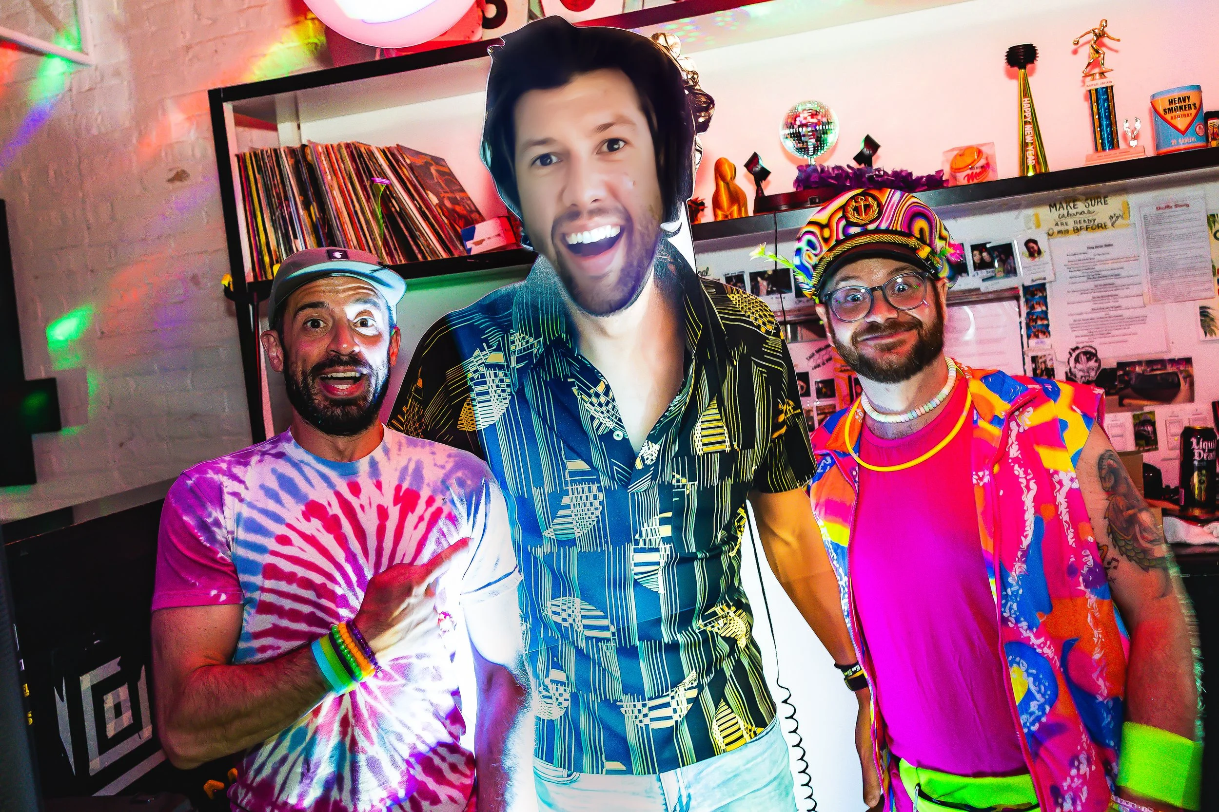 Three men at a colorful party, wearing vibrant, retro-style clothing and accessories, smiling and posing for the camera.