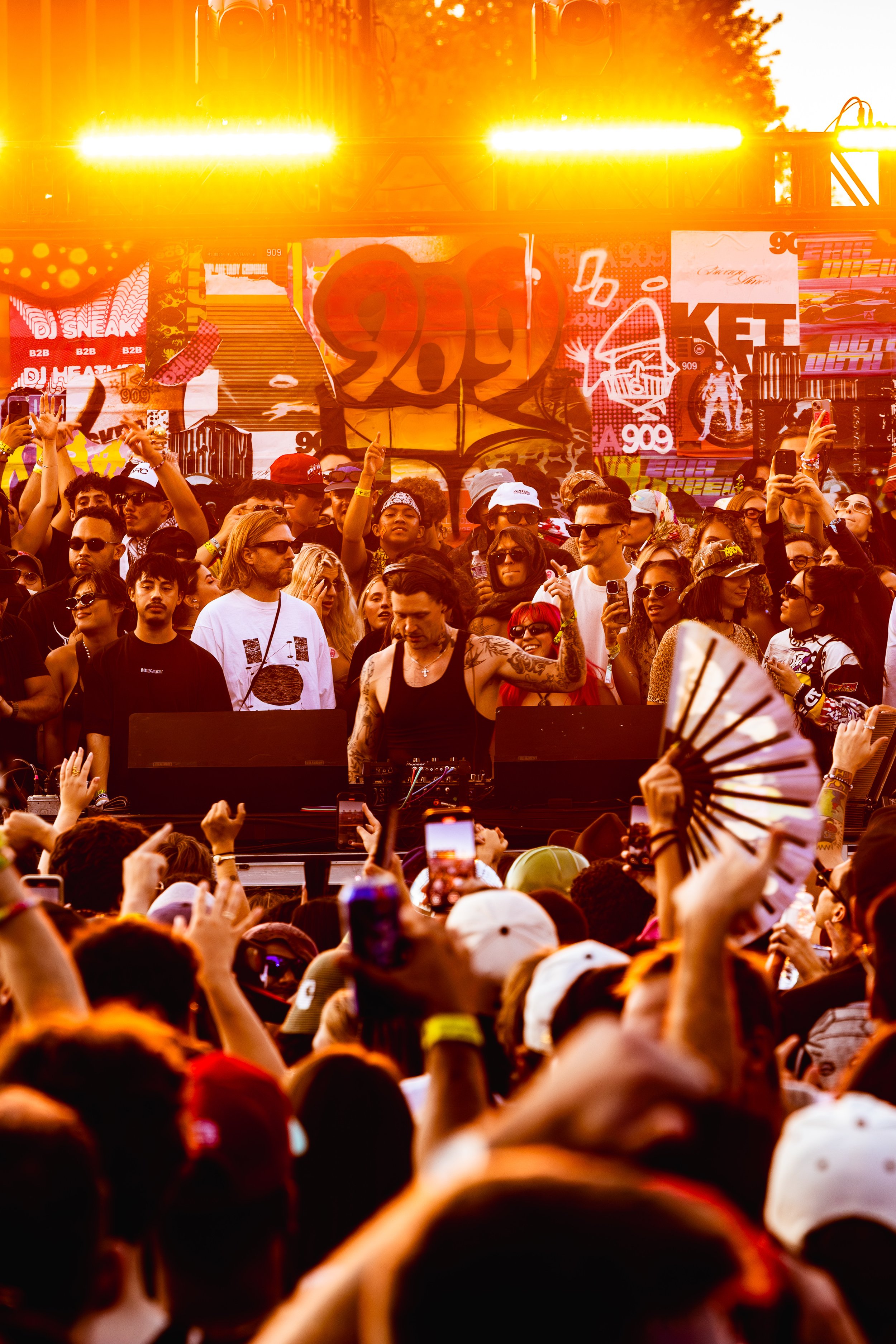 A scene at an outdoor music festival with a DJ playing in front of a large crowd, colorful banners, and vibrant lighting.