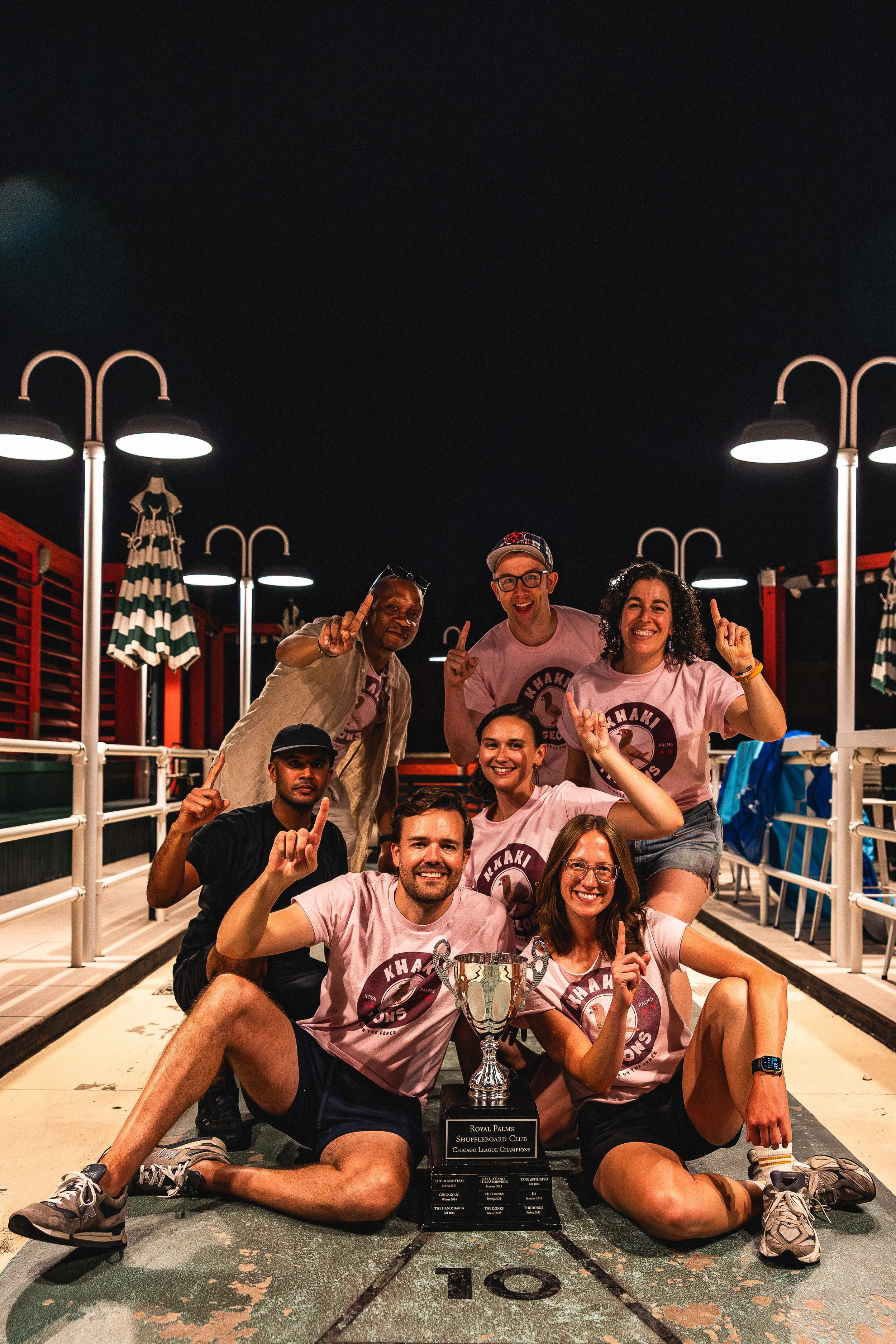 Group of people celebrating a victory at a shuffleboard court at night, holding a trophy, all raising their index fingers, some wearing matching pink t-shirts with a shuffleboard logo, smiling and posing.