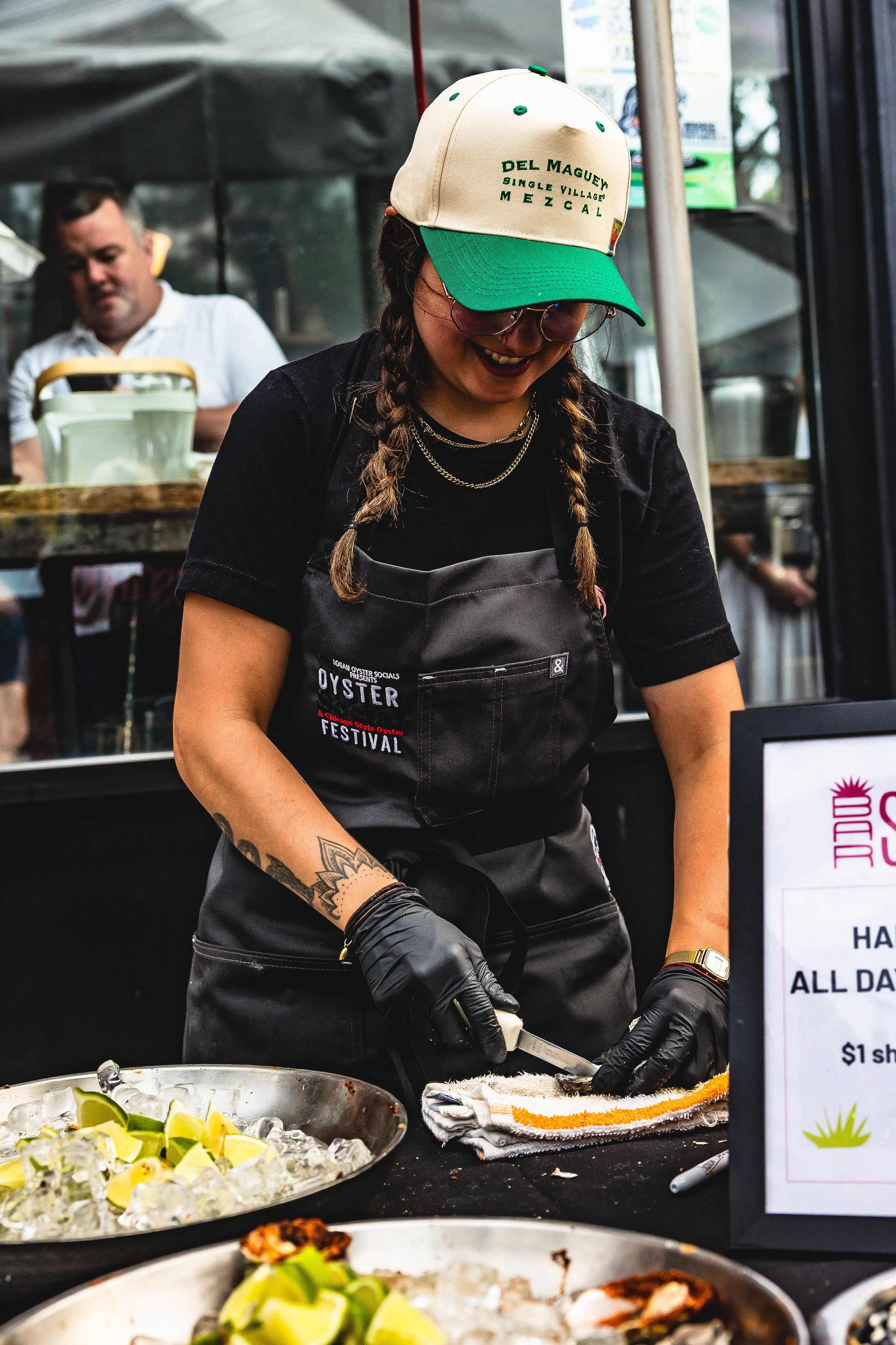 Woman preparing food at a food stall, wearing a green and white cap, black apron, and black gloves, with a man in the background.