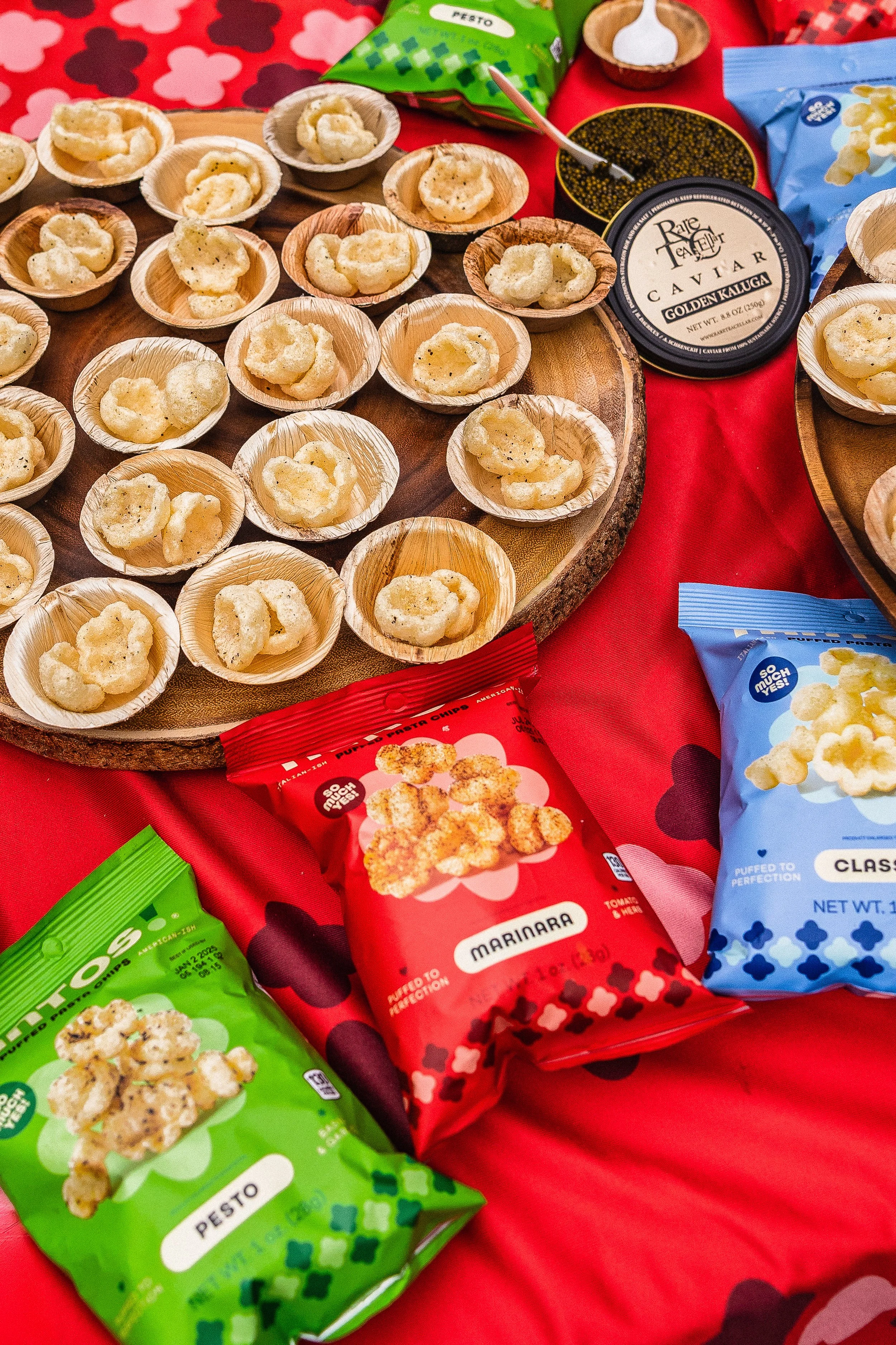 Assorted flavored popcorn bags and small bowls of popcorn on a wooden tray set on a red tablecloth with black and red heart patterns.