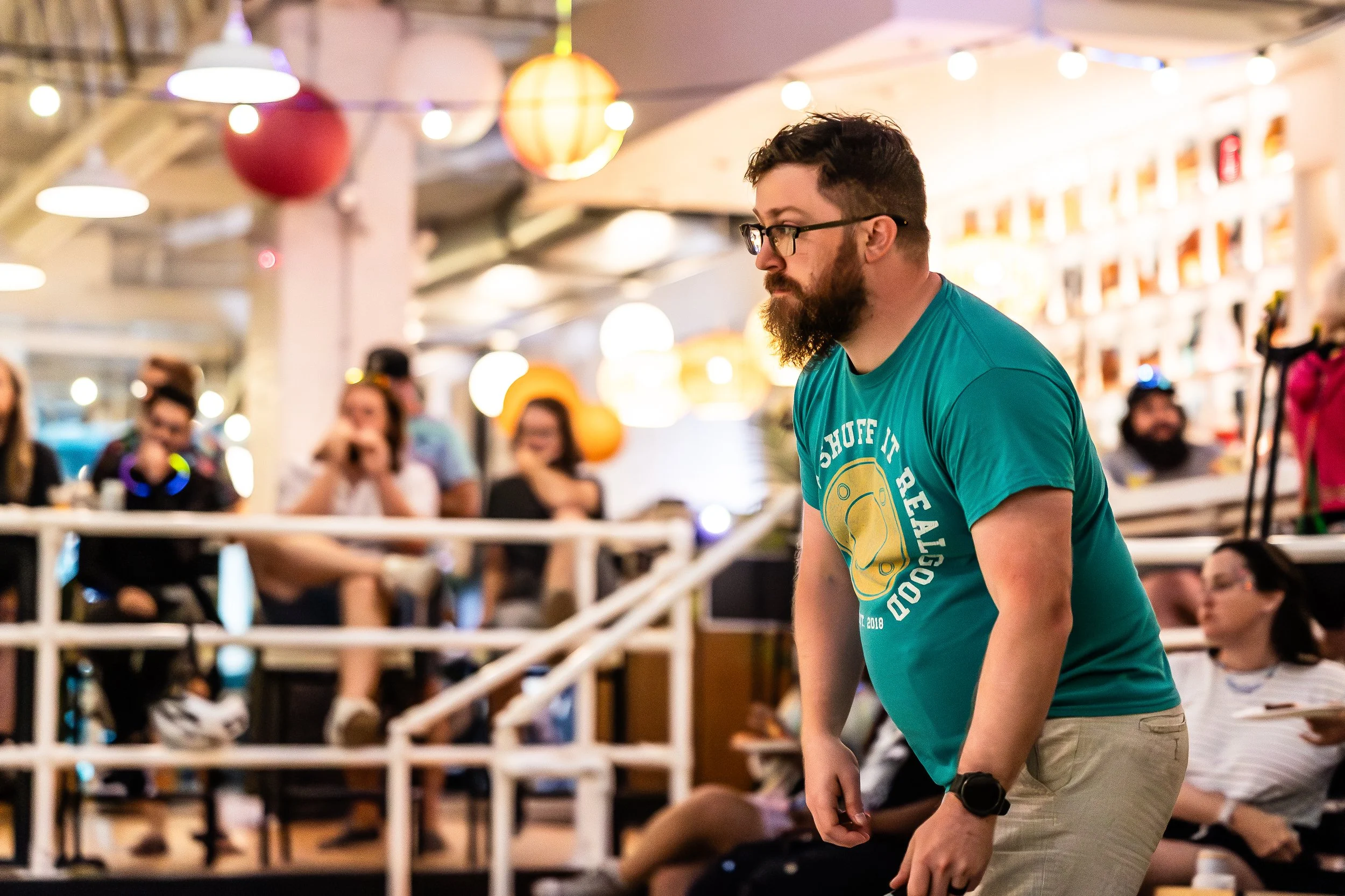 A man with a beard and glasses in a teal T-shirt and khaki pants is in a crowded indoor space, possibly a game or competition, surrounded by seated and standing people, with colorful lanterns hanging from the ceiling.