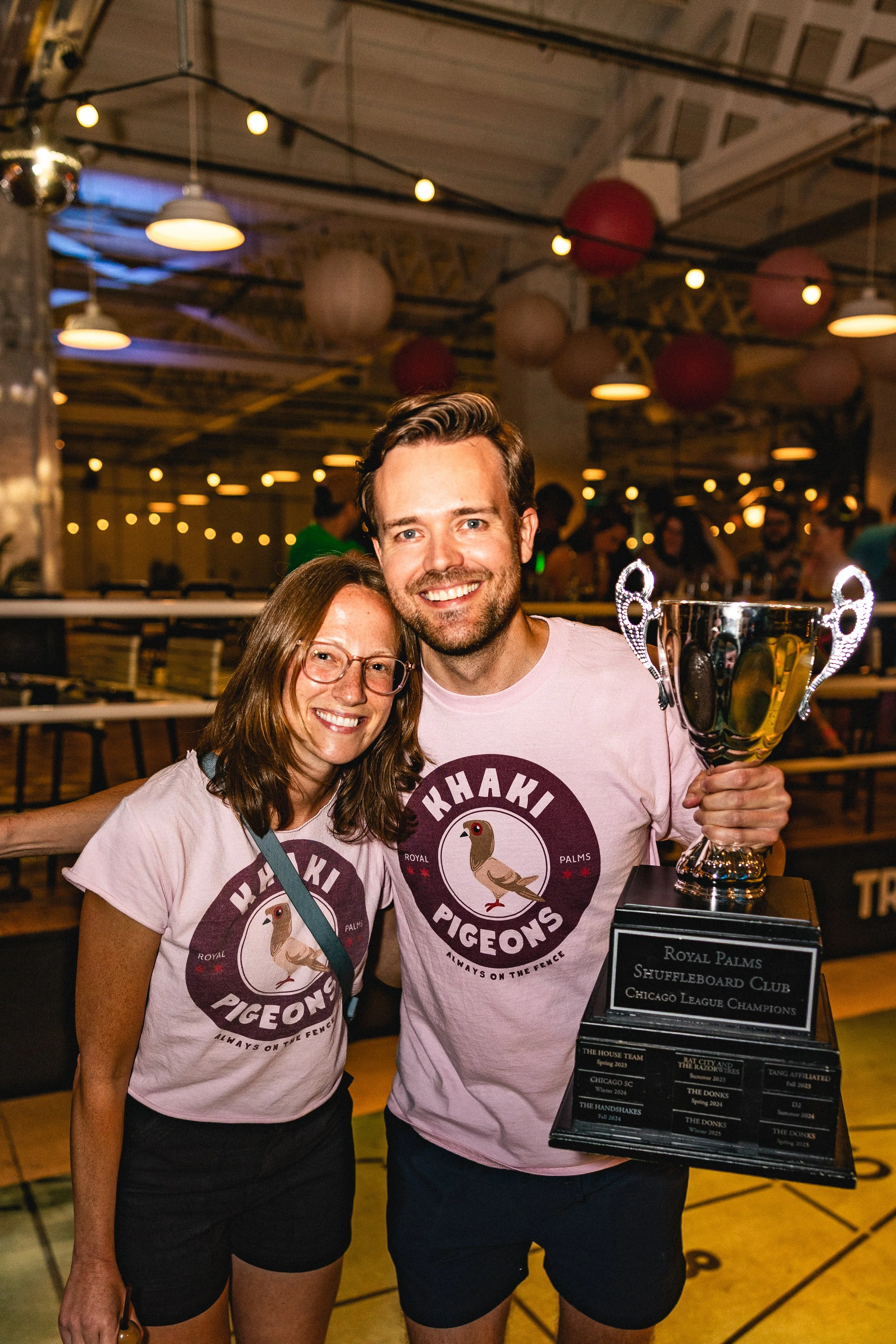 Two people are celebrating, holding a trophy and a plaque at an indoor event, both wearing matching pink T-shirts with a pigeon logo and the text 'KHAKI PIGEONS ALWAYS ON THE FENCE', in a decorated space with hanging lights and balloons.
