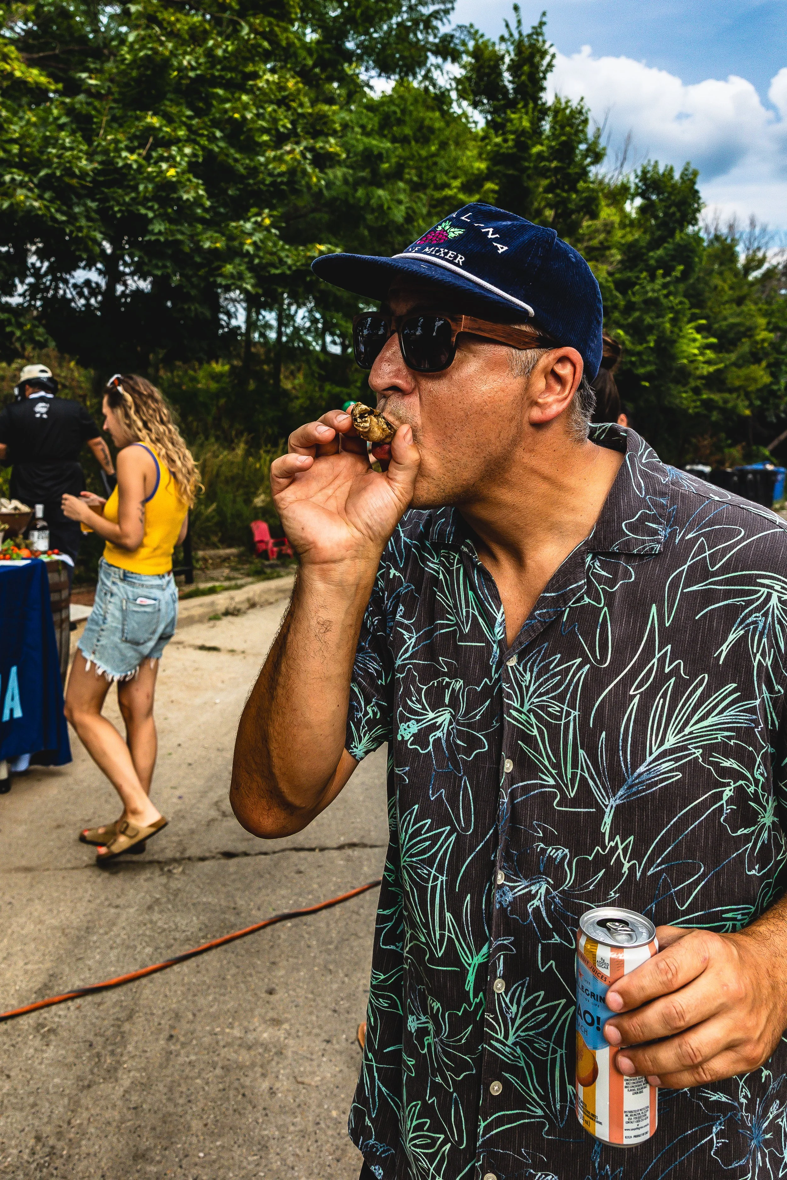Man wearing sunglasses, a blue cap, and a Hawaiian shirt eating a grilled oyster at an outdoor gathering, holding a can of beer.