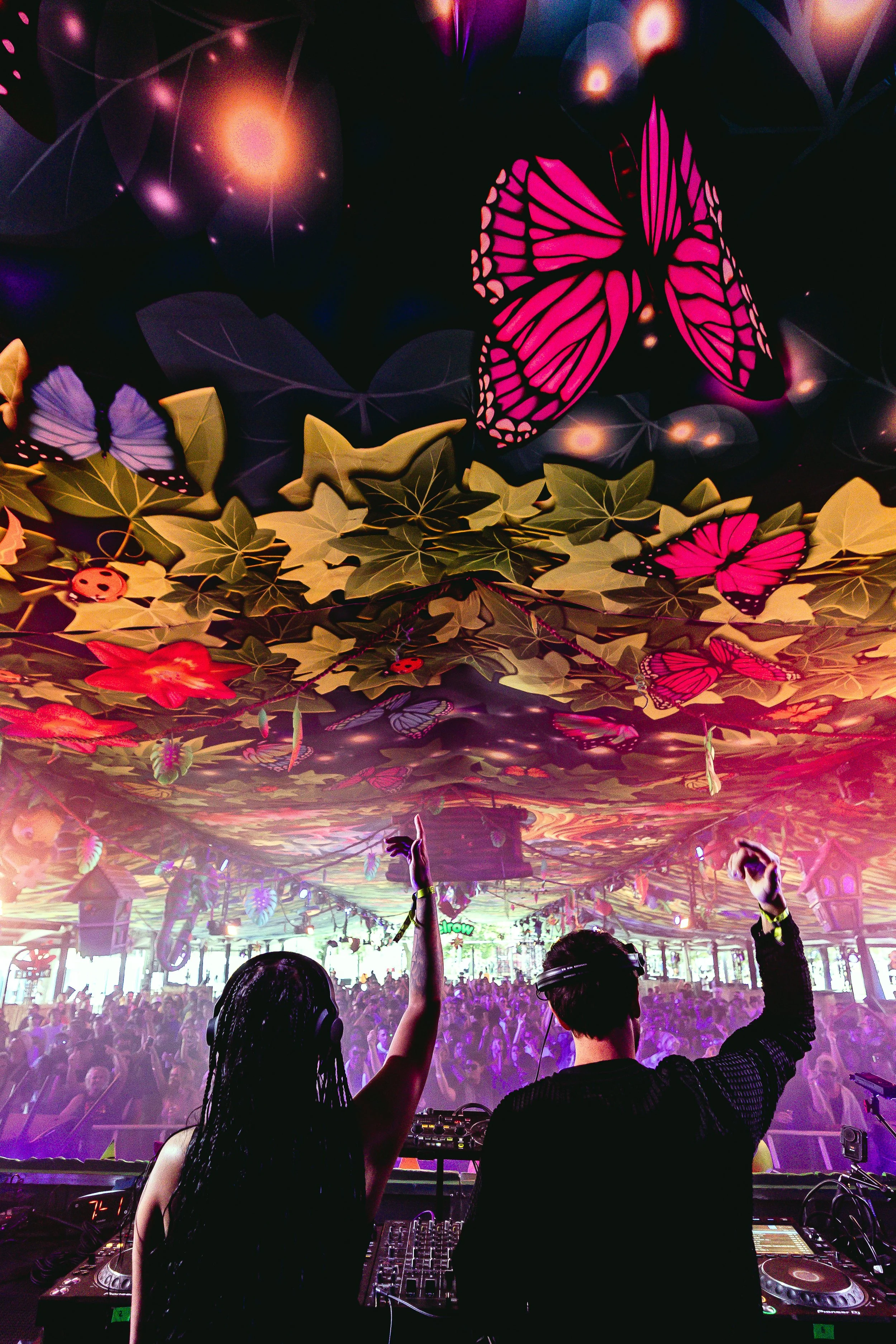 A crowded indoor music event with two DJs performing at the front, colorful ceiling decorations featuring large pink butterflies and leafy patterns, and a lively audience dancing under vibrant lighting.