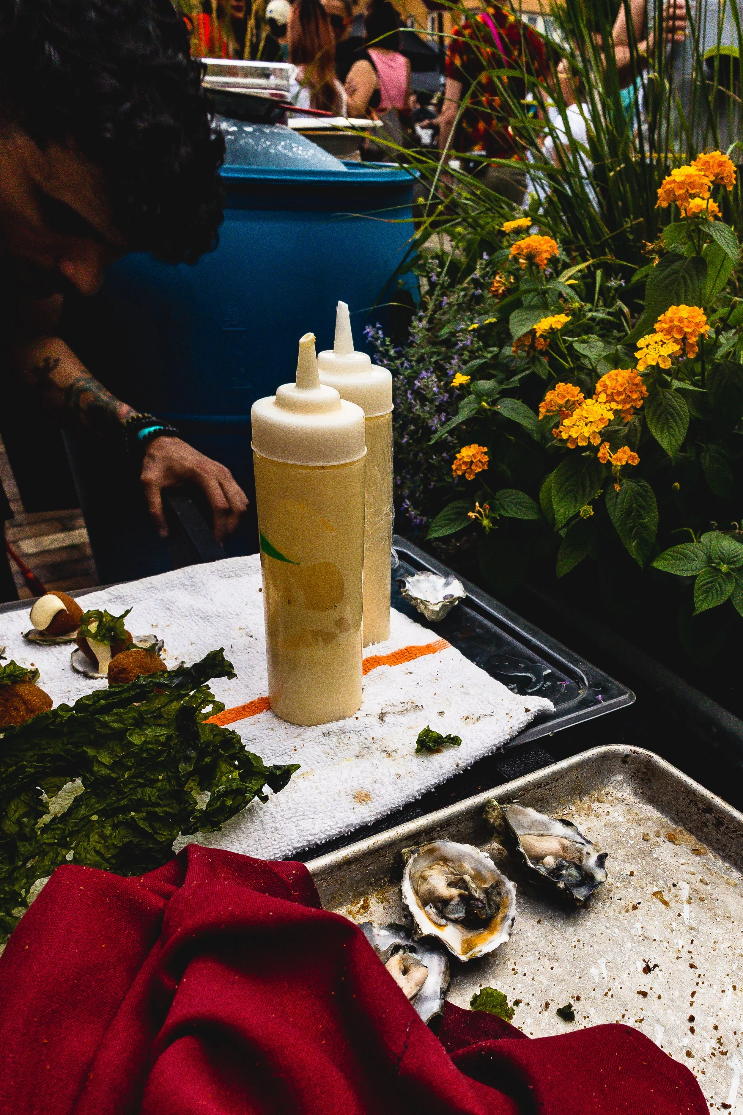 A food stall at an outdoor market with two bottles of condiments, oysters, greens, and flowers in the background, with people walking by.
