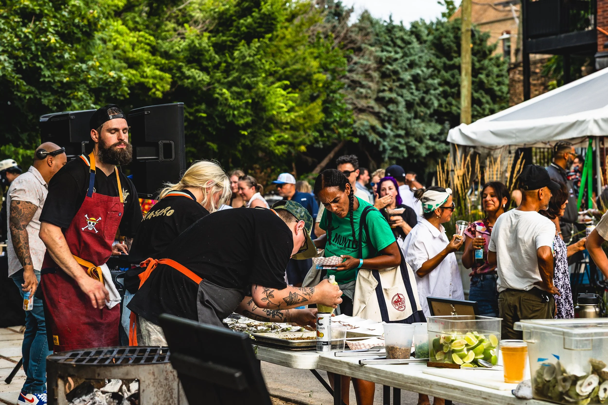 People gathered at an outdoor event, some wearing aprons and cooking, with food and drinks on tables, surrounded by trees and a tent, indicating a food festival or cookout.