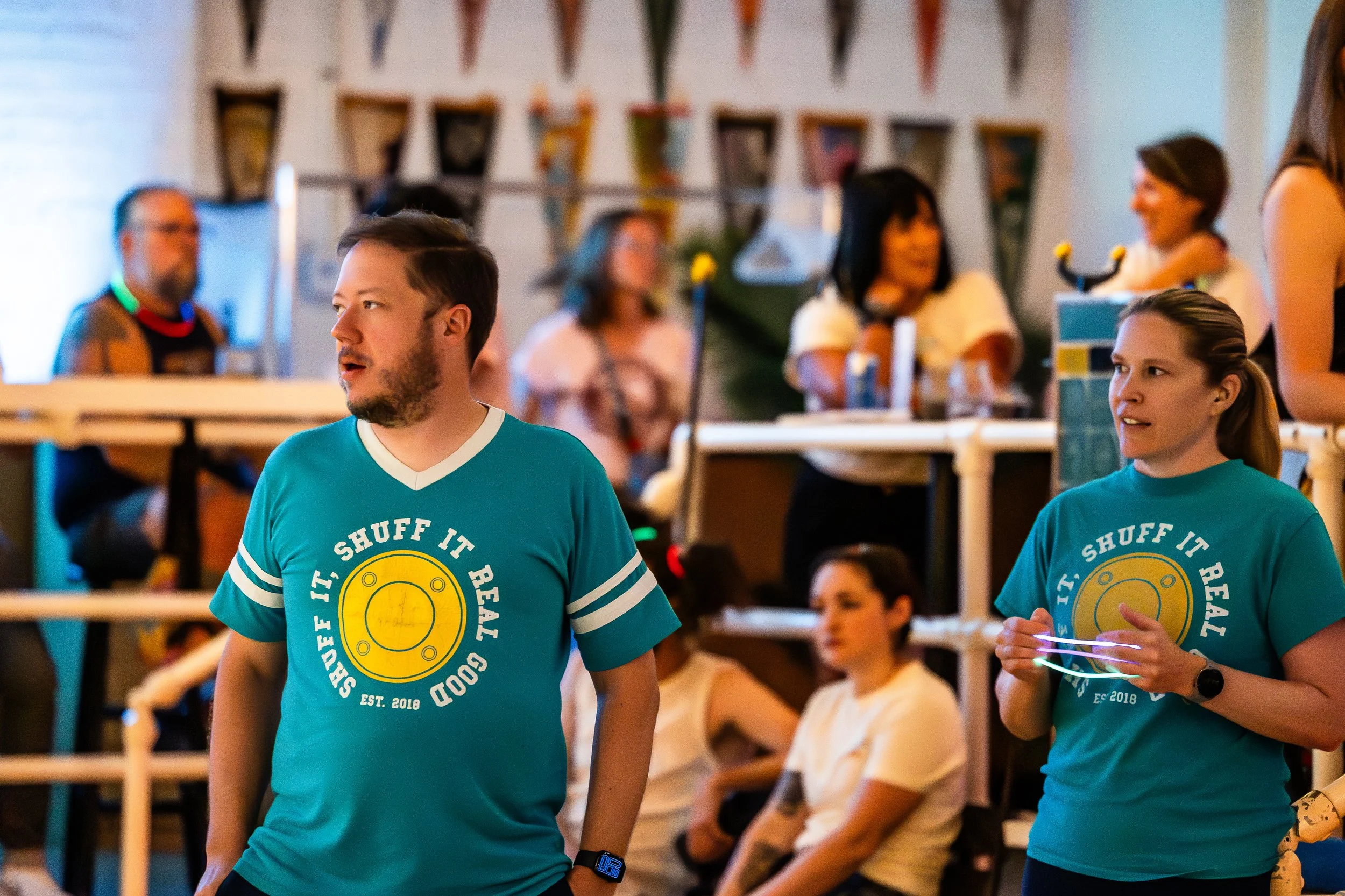 People at a social gathering, some wearing matching teal t-shirts with the phrase 'SHUFF IT, SHUFF IT, REAL GOOD' and a graphic of a circular object, in a brightly decorated indoor space.