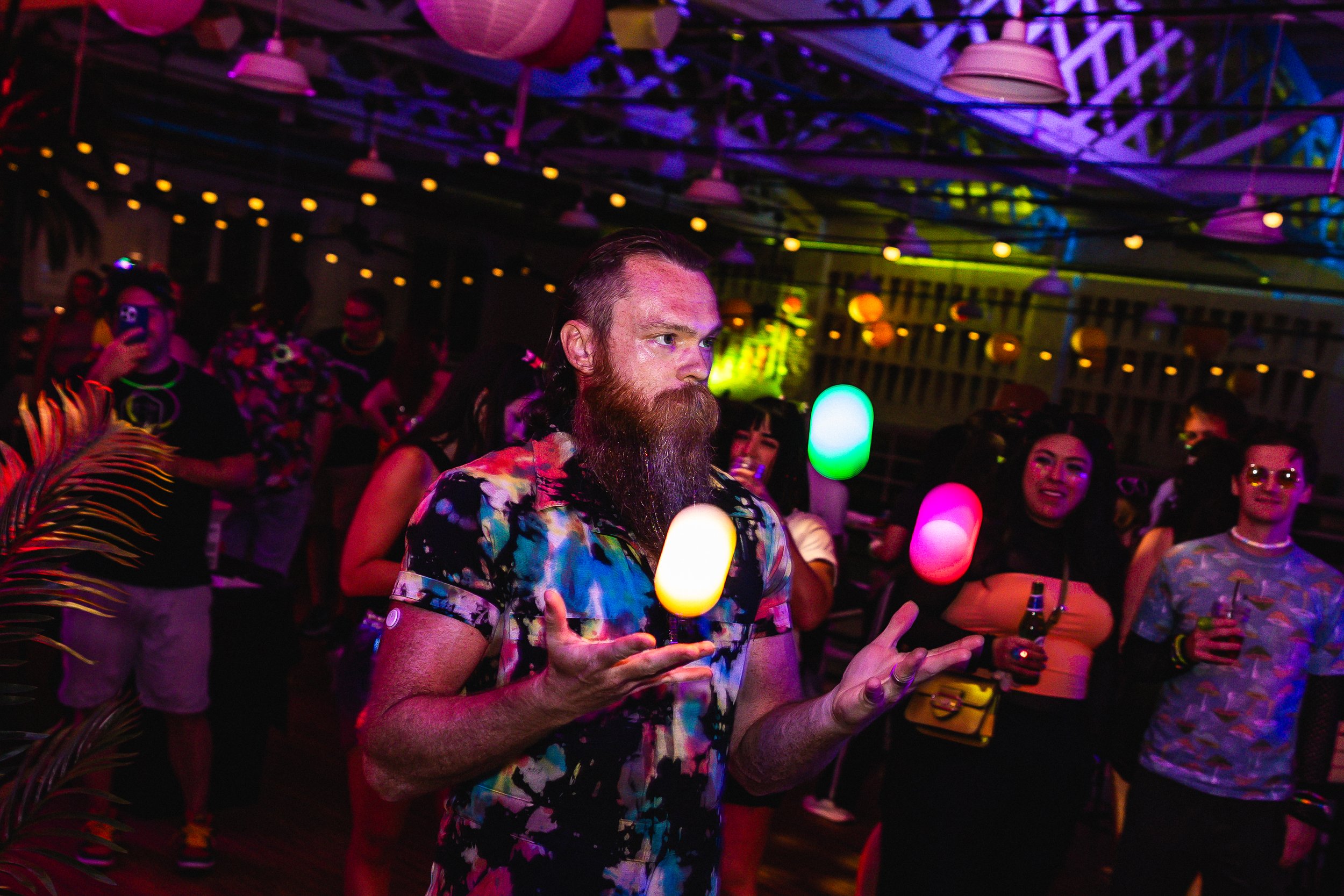A man with a beard and tattooed arms juggling colorful LED lights at a lively indoor party with people dancing and socializing under colorful lighting.