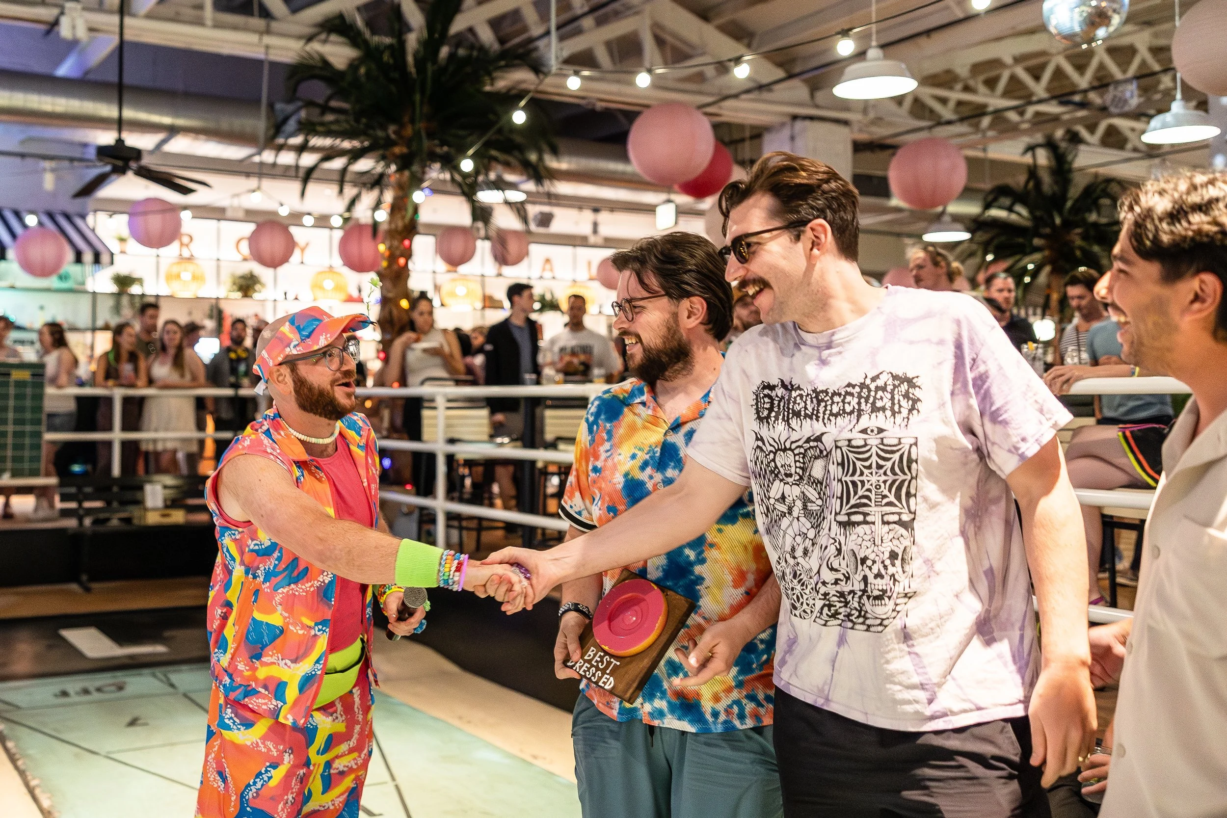 People smiling and shaking hands at a lively indoor party or event, decorated with pink paper lanterns, plants, and string lights, with many onlookers in the background.