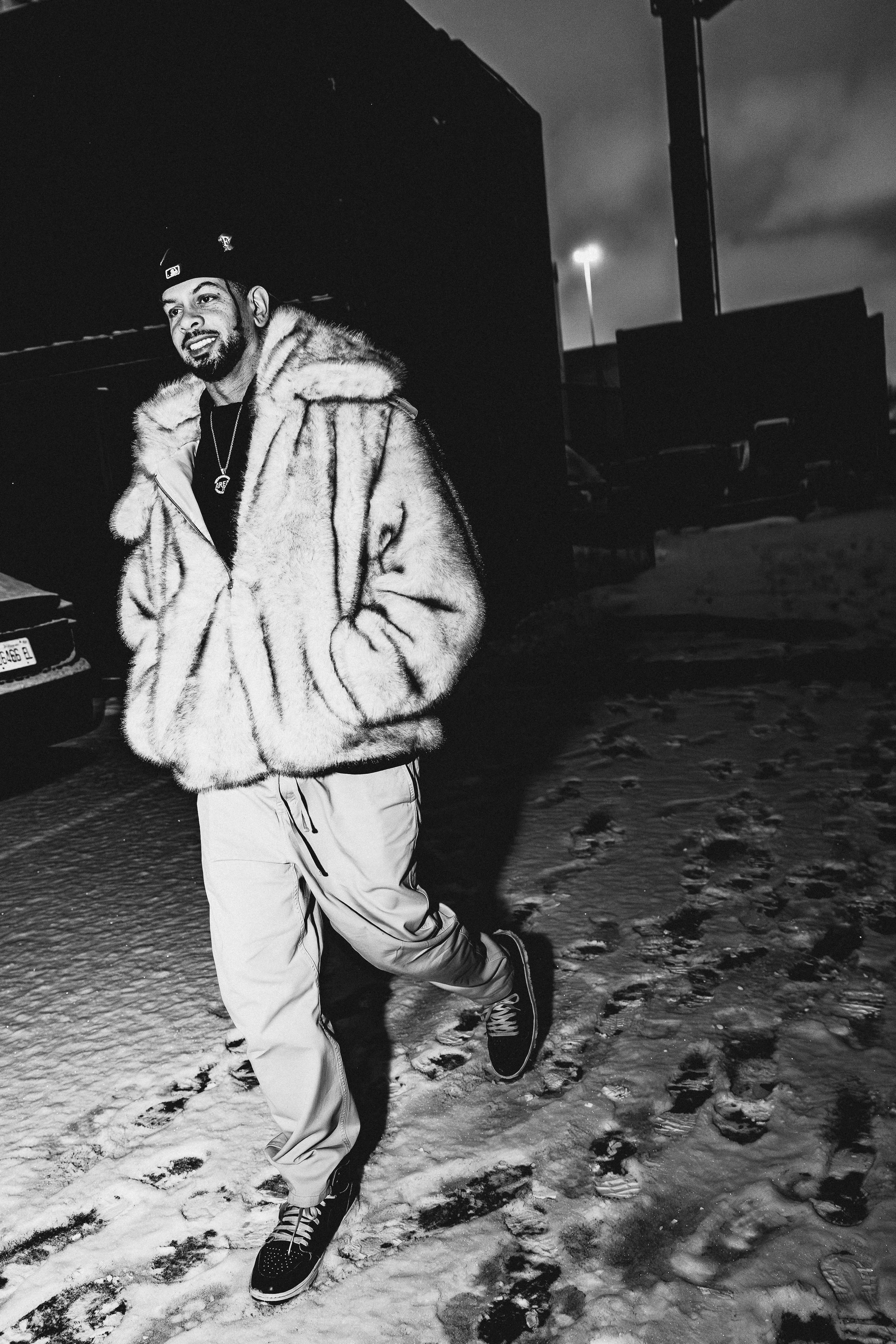 Marc Kinchen, known by his initials MK, walking outdoors on snowy ground at night, wearing a fur coat, light-colored pants, and sneakers.