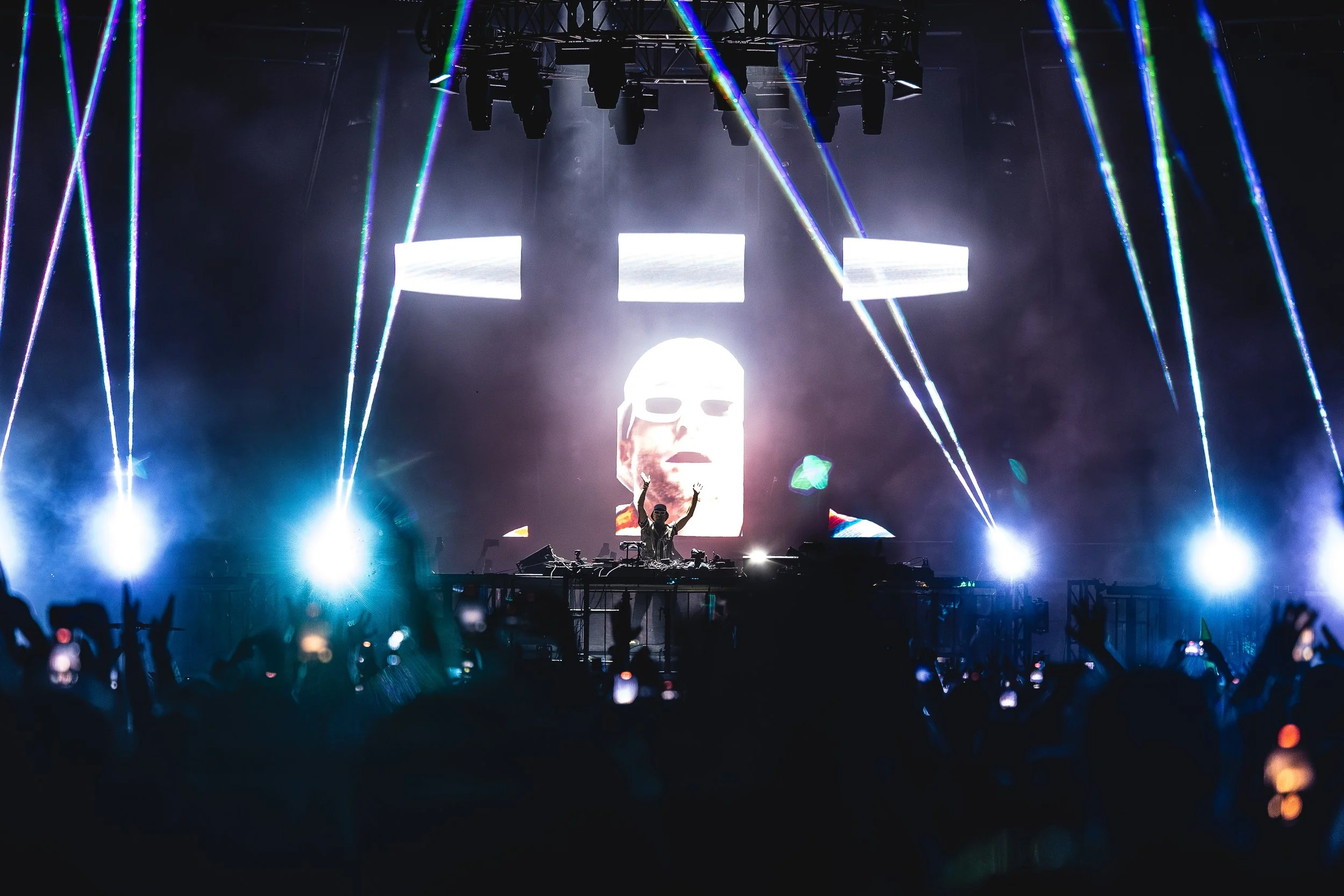 A DJ performing on stage at a concert with colorful laser lights and a large screen displaying a face wearing sunglasses behind him.
