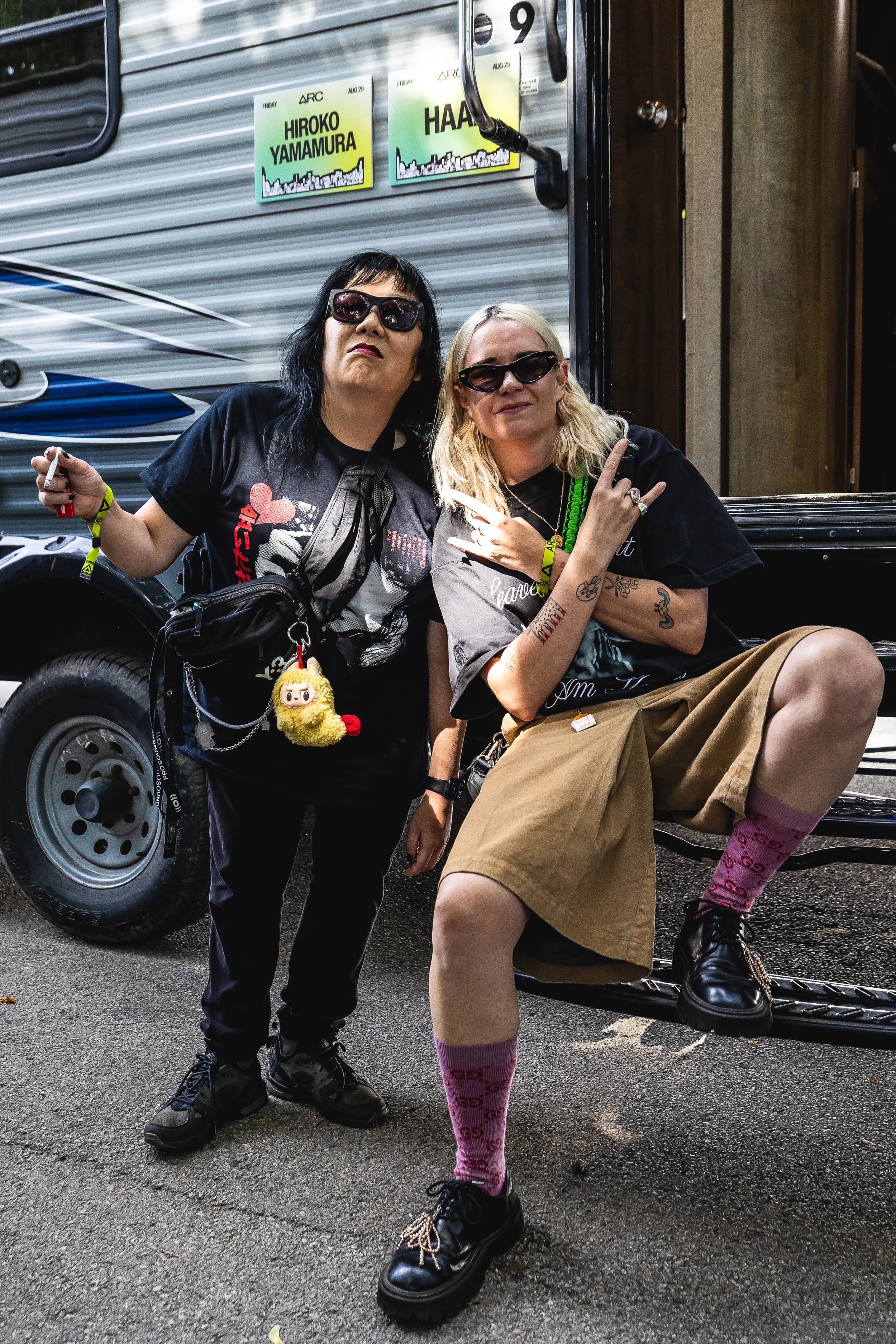 Two women posing in front of a vehicle at an outdoor event, with one sitting on the vehicle and the other standing beside it. They are wearing sunglasses and casual clothing.