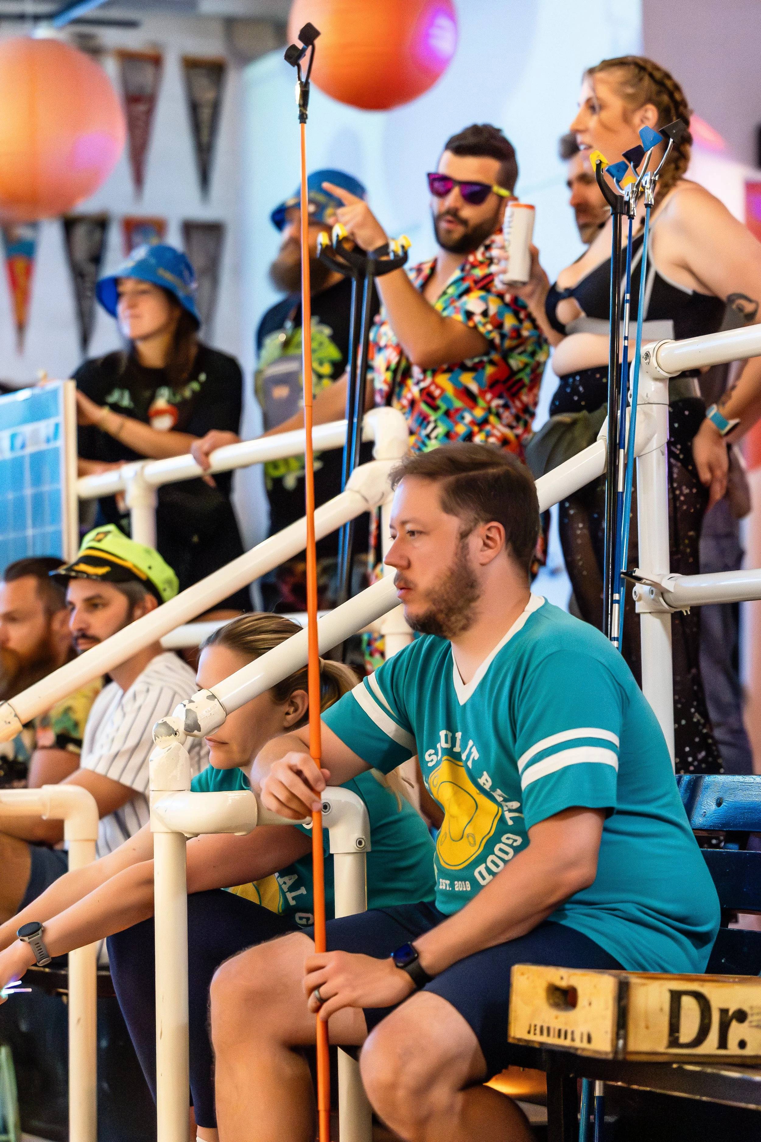 Group of people at a party or event, some sitting and some standing, with colorful decorations and lighting, wearing casual and festive clothing.