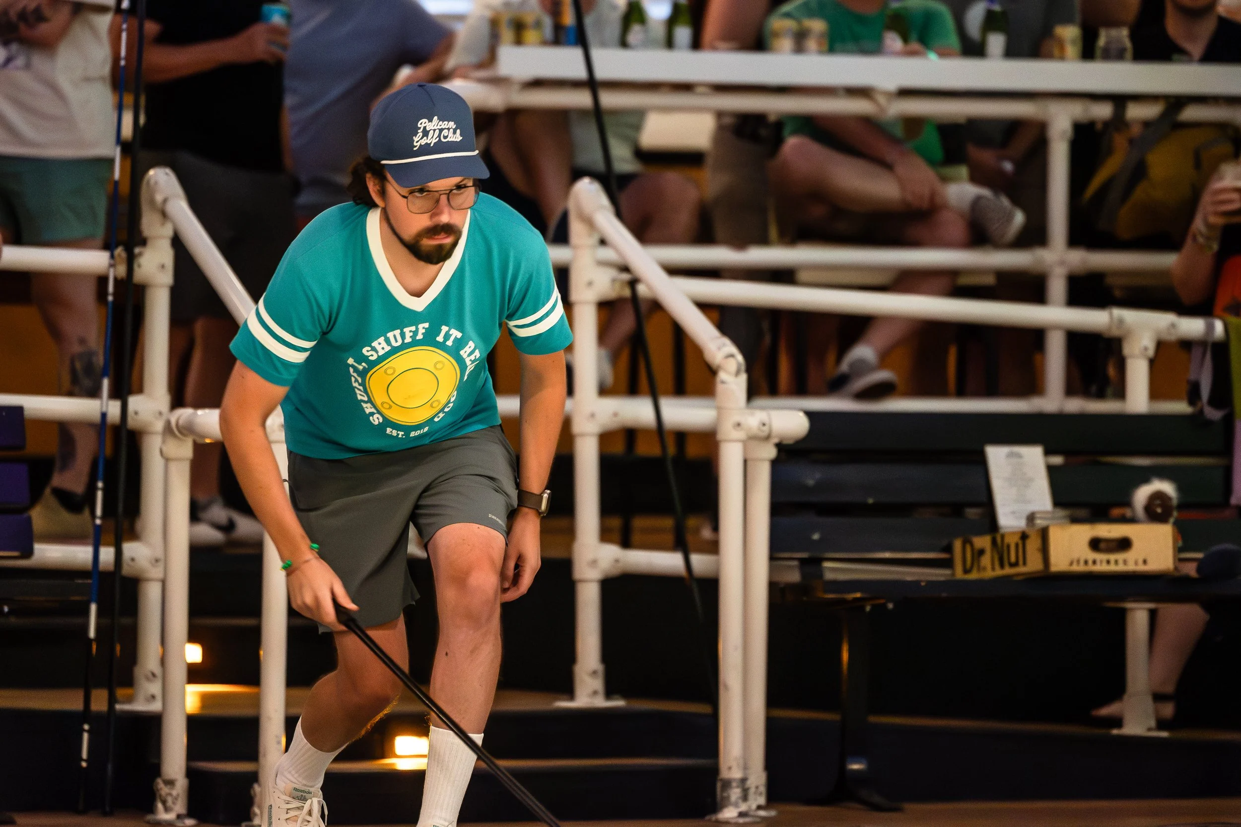 A man wearing a teal T-shirt with a yellow disc design and the words 'Shuff It, Shuffle It, Game On', a navy cap, glasses, gray shorts, and white sneakers is participating in a shuffleboard game indoors, with spectators seated in the background.