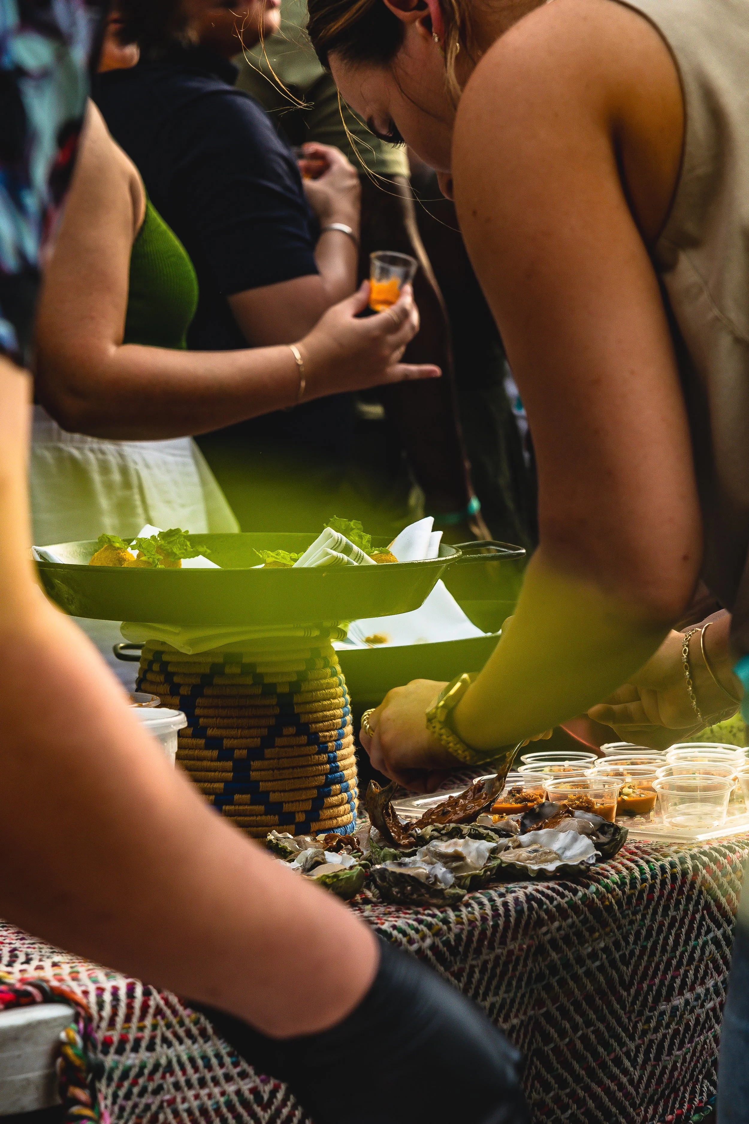 People gathered around a table at a food event, serving and enjoying dishes. The table is decorated with a colorful, woven cloth, and food trays and small cups are visible.