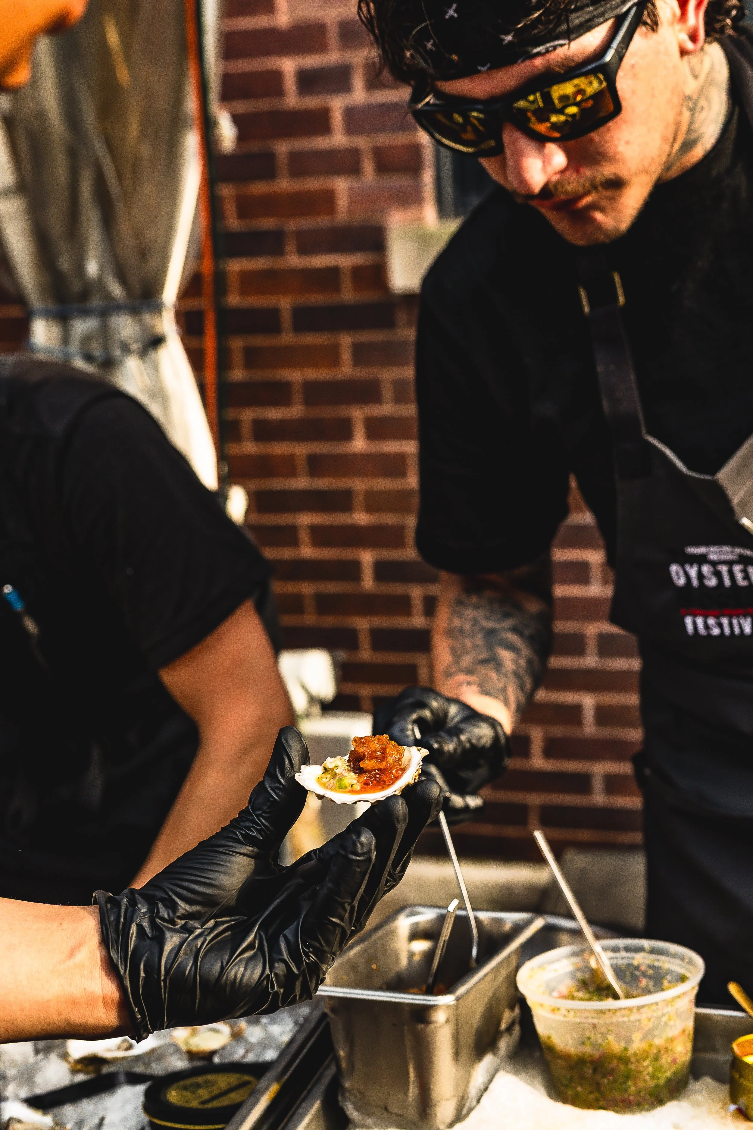 A person wearing black gloves holds a small oyster shell with a topping, while another person wearing sunglasses and a bandana leans over a table with food and utensils, set against a brick wall.