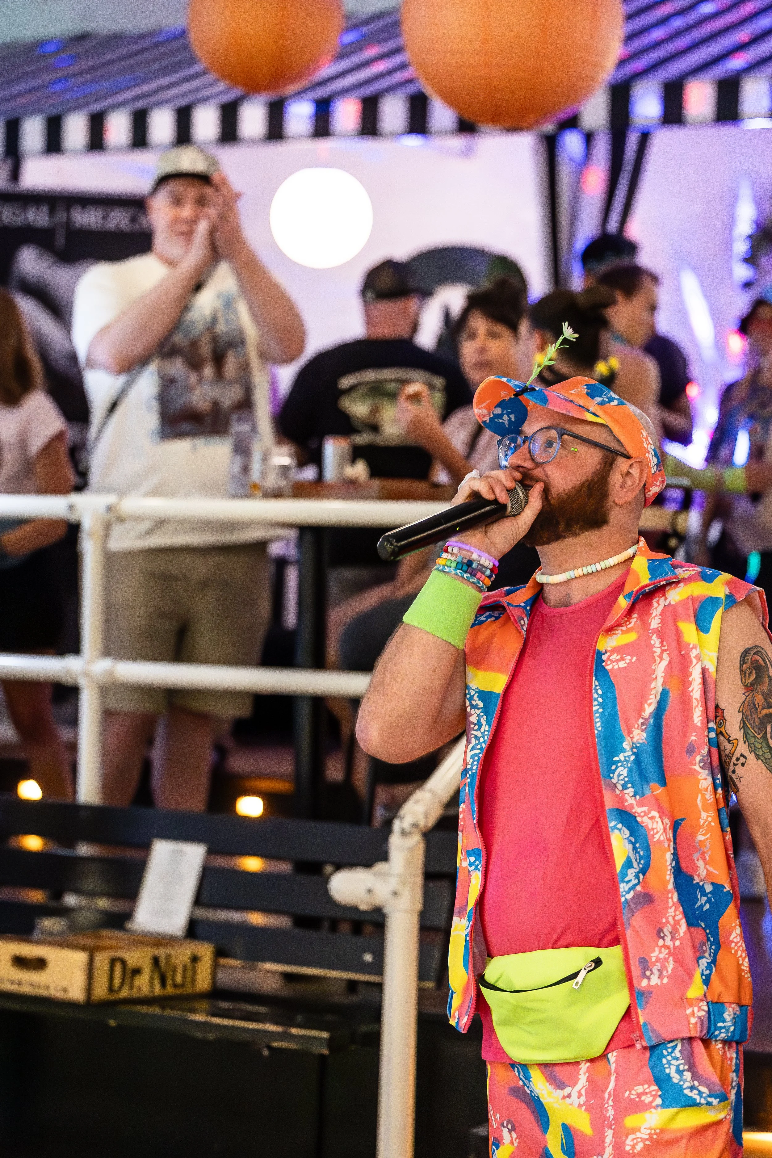 A performer dressed in a colorful, tropical-themed outfit, wearing a cap with a plant sprout, glasses, and bracelets, holding a microphone at an event with a lively crowd in the background.