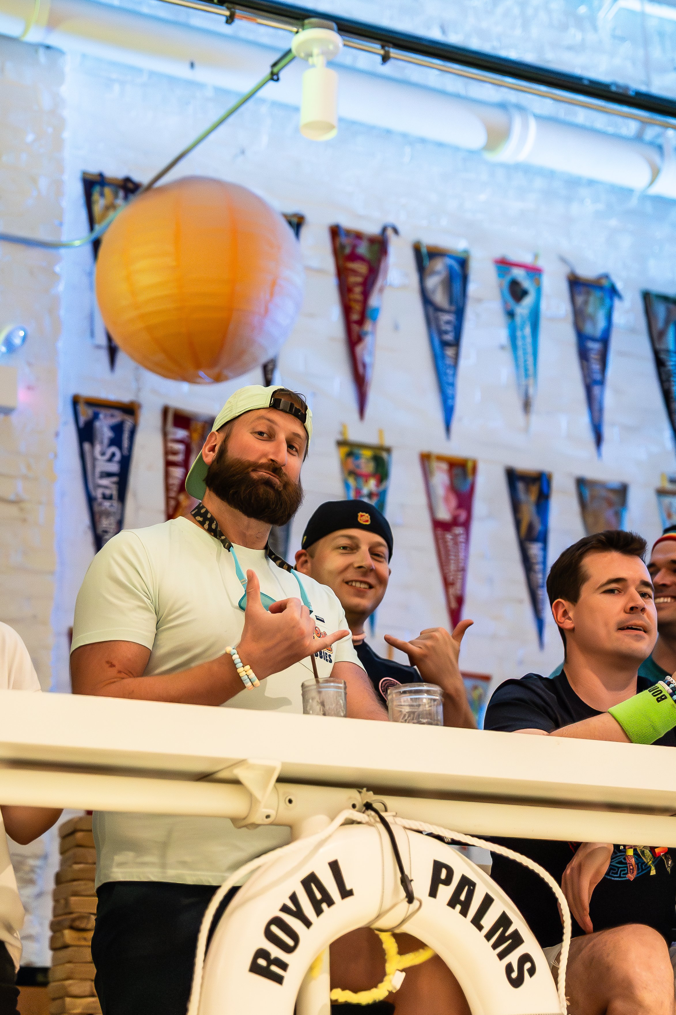 Three men sitting behind a boat labeled 'Royal Palms,' with colorful pennant banners hanging on a white brick wall in the background. One man with a beard is wearing a white t-shirt and a backwards cap, making a hang loose gesture with his hand. The 