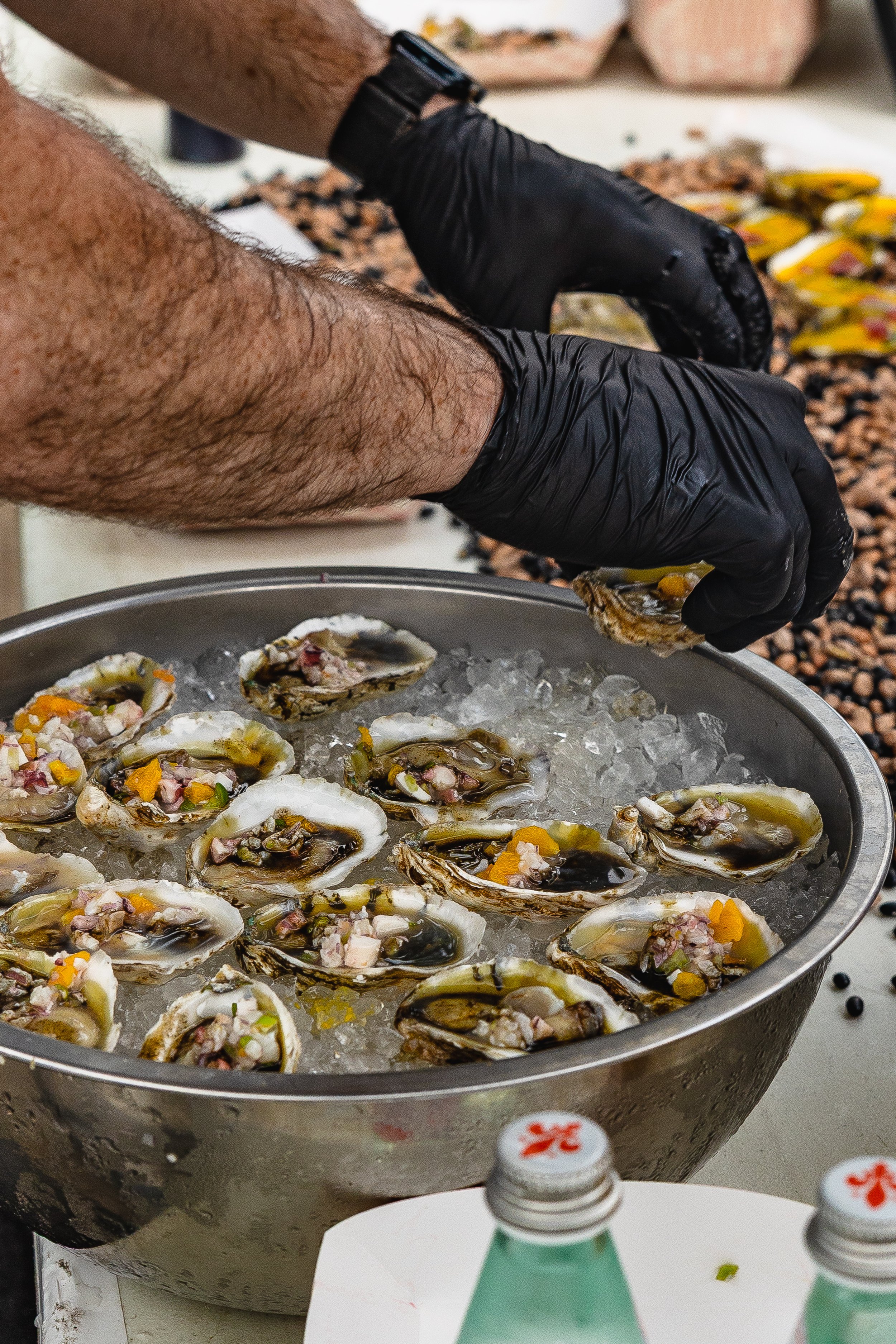 Person wearing black gloves preparing oysters on a bed of ice, with a background of spilled beans and banana peppers.