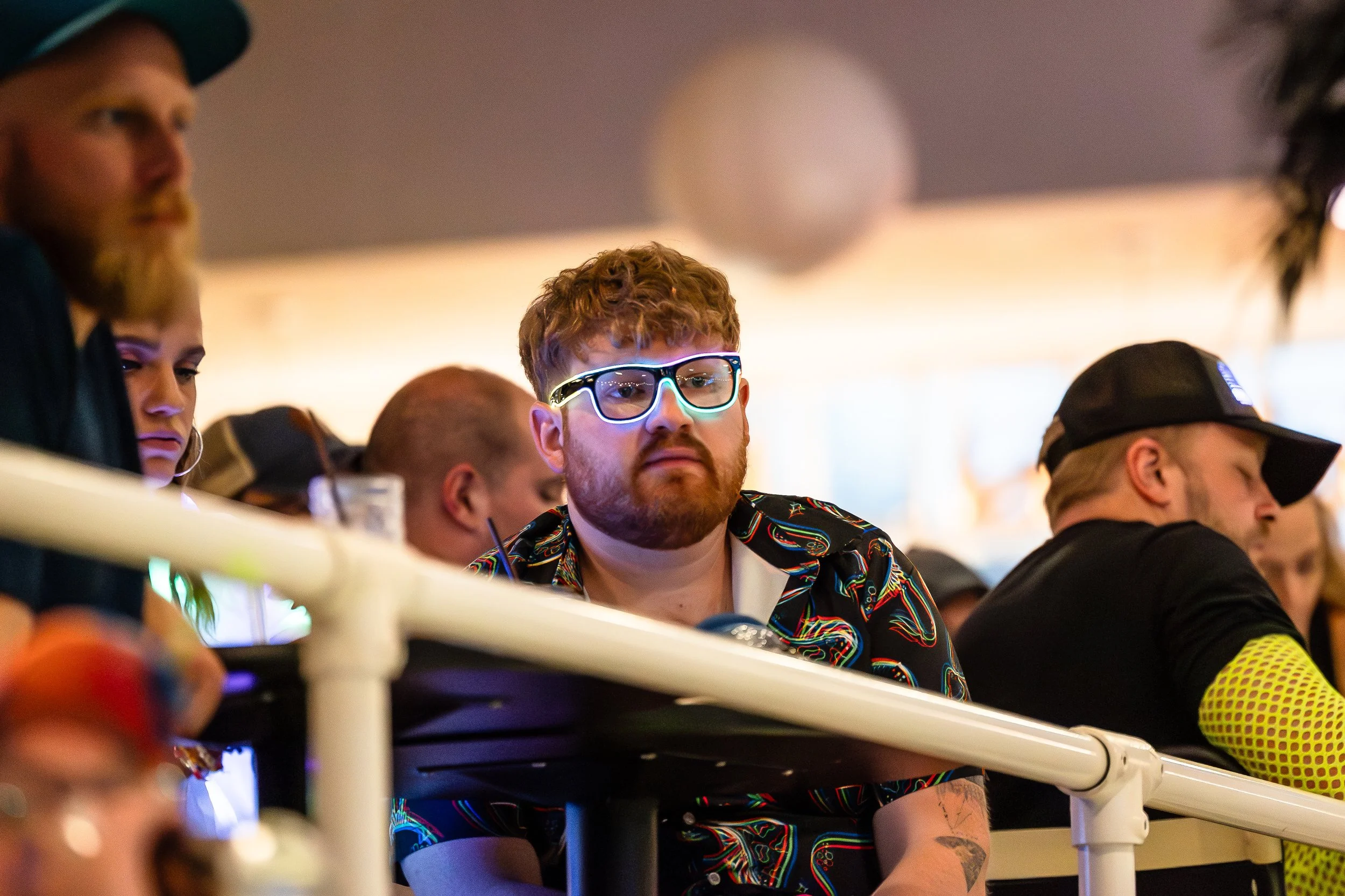 A man with curly hair and a beard wearing glasses and a colorful shirt, sitting among other people in an indoor setting, looking focused.